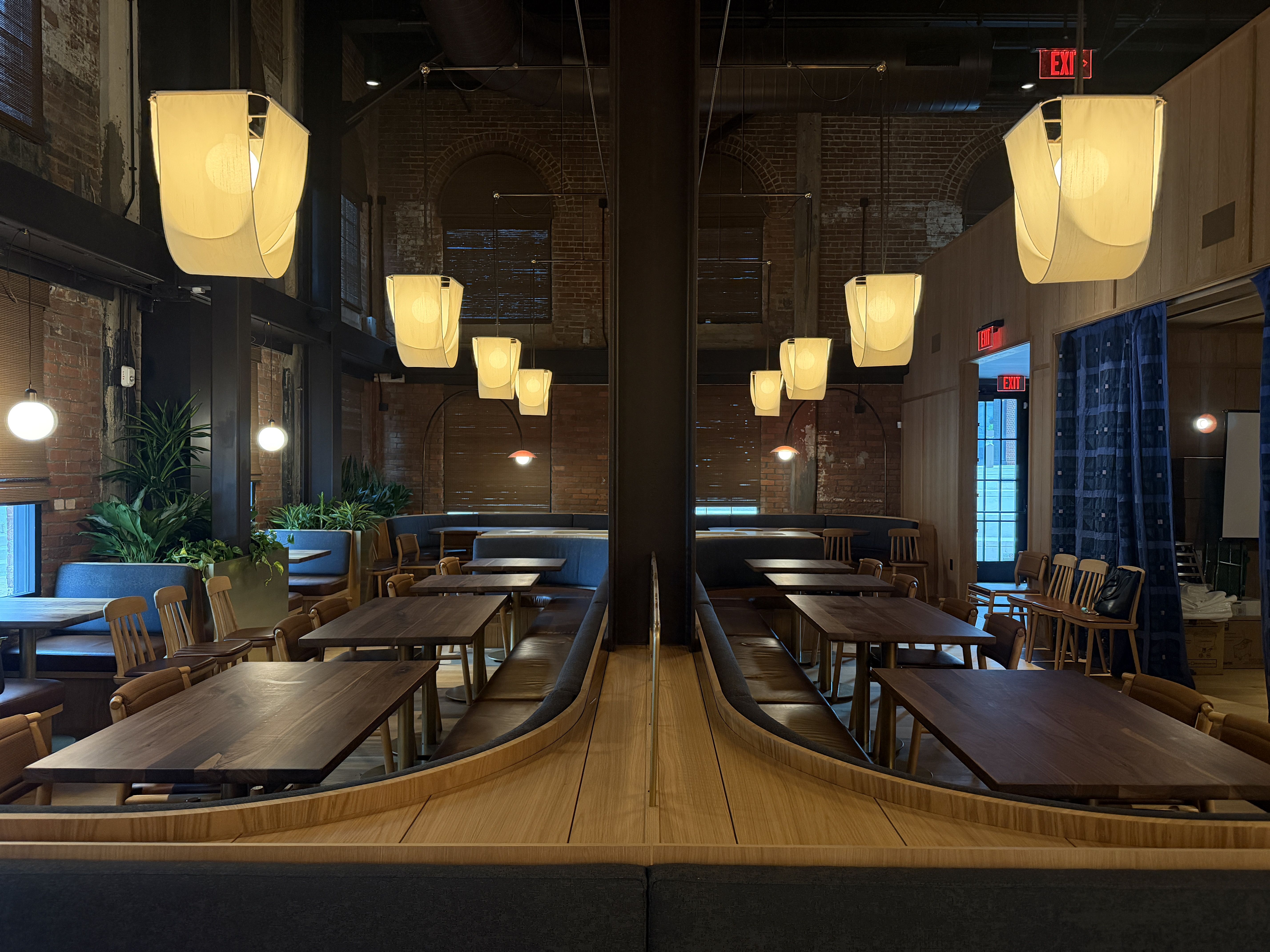 Interior of a cozy restaurant with wooden tables and chairs, soft hanging yellow lamps, green plants, exposed brick walls, and a large mirror reflecting the seating area symmetrically.