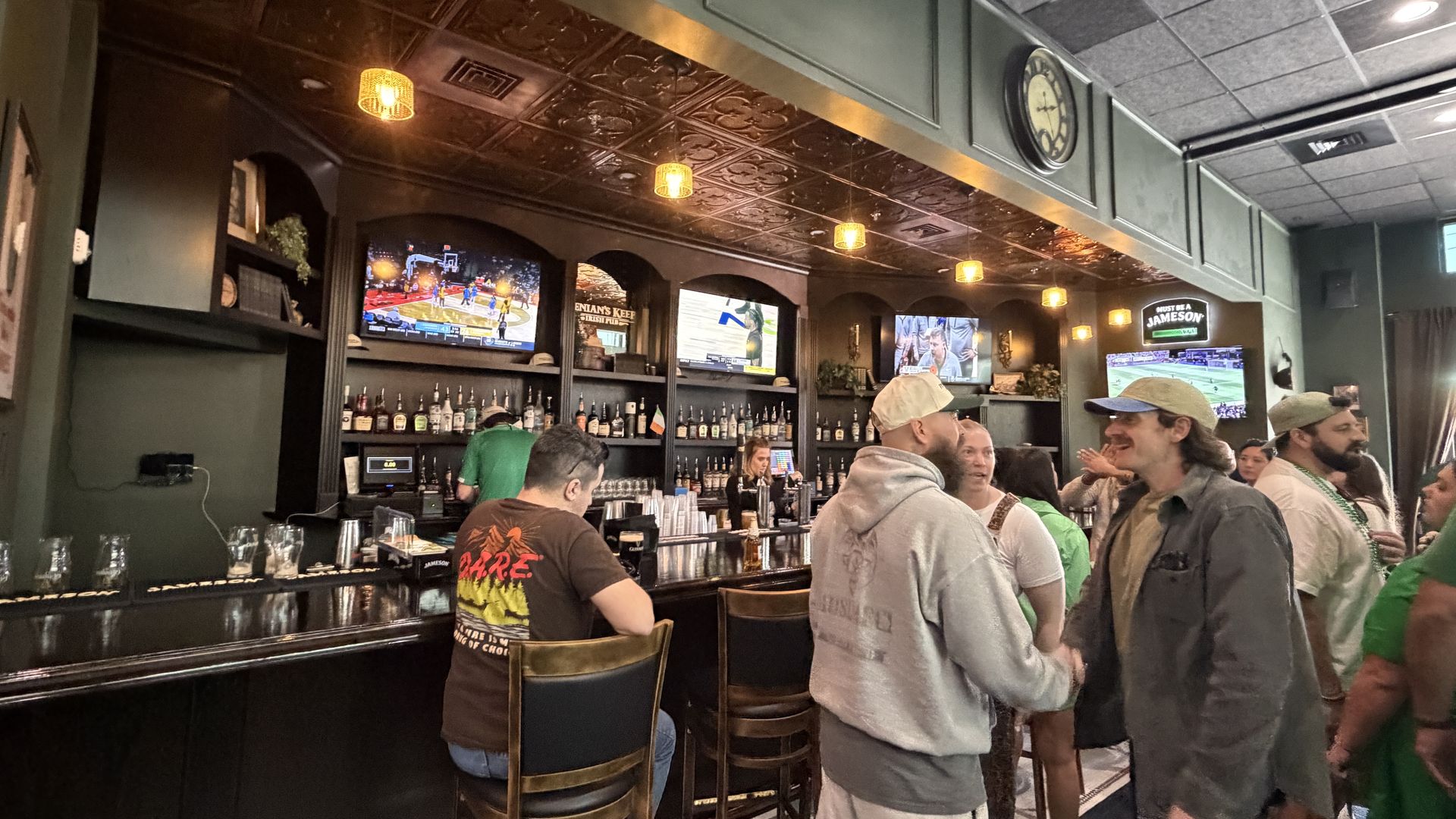 A crowded sports bar with dark wood shelves and bottled drinks behind a long black counter. Patrons chat, some shake hands near stools as TVs show basketball and football; warm pendant lights glow.