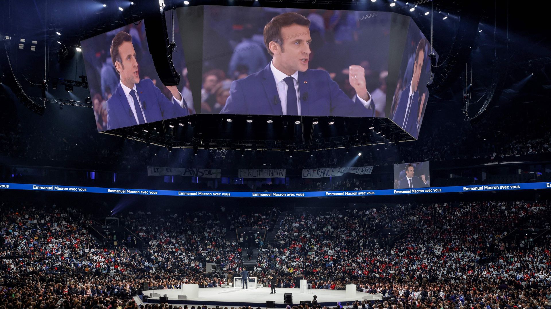 Macron speaking on a jumbotron in an arena.