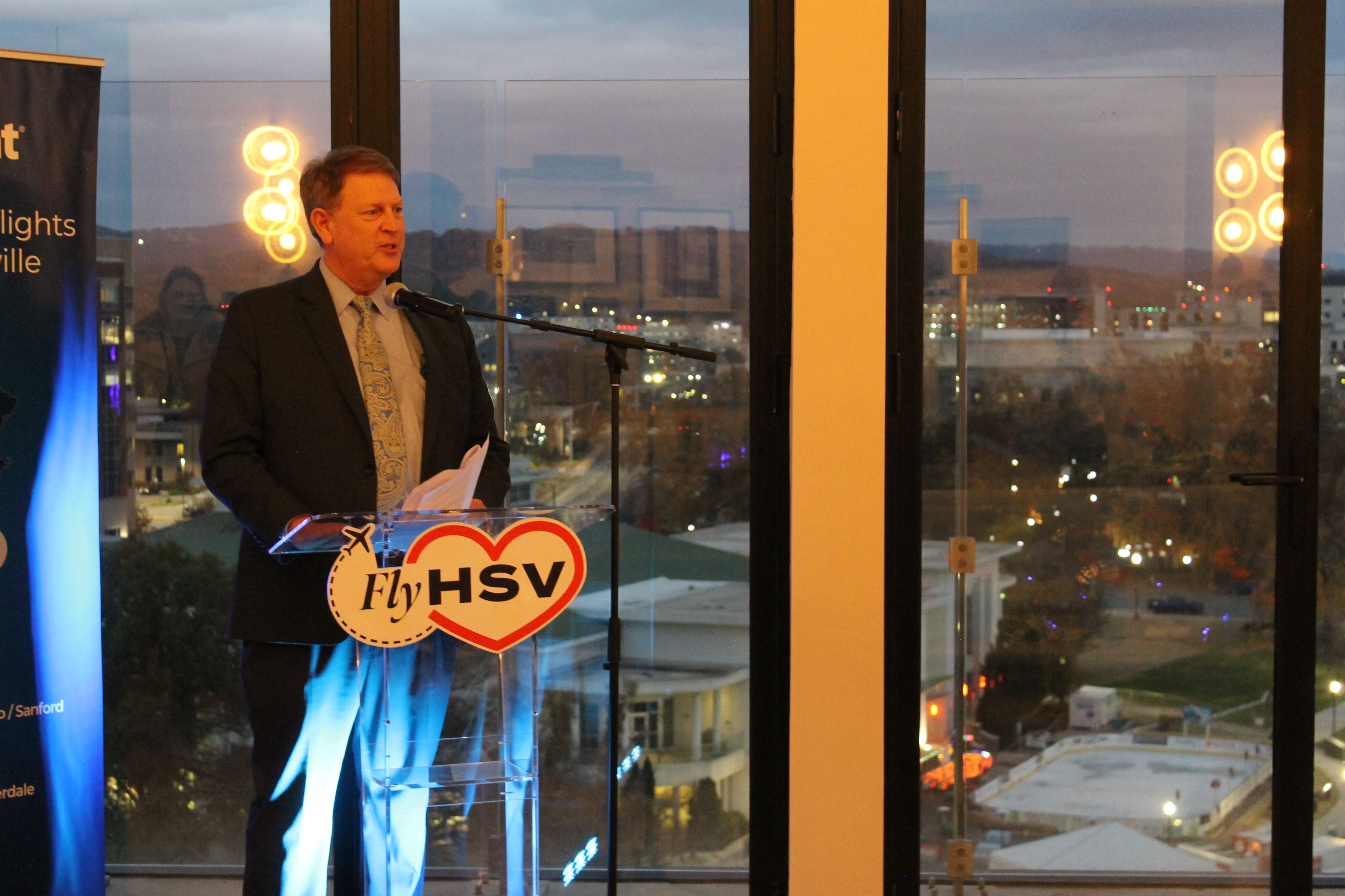 Man in a dark suit and paisley tie speaking at a clear podium with a "Fly HSV" logo, in front of large glass windows showing a cityscape at dusk with lights and buildings in the background.