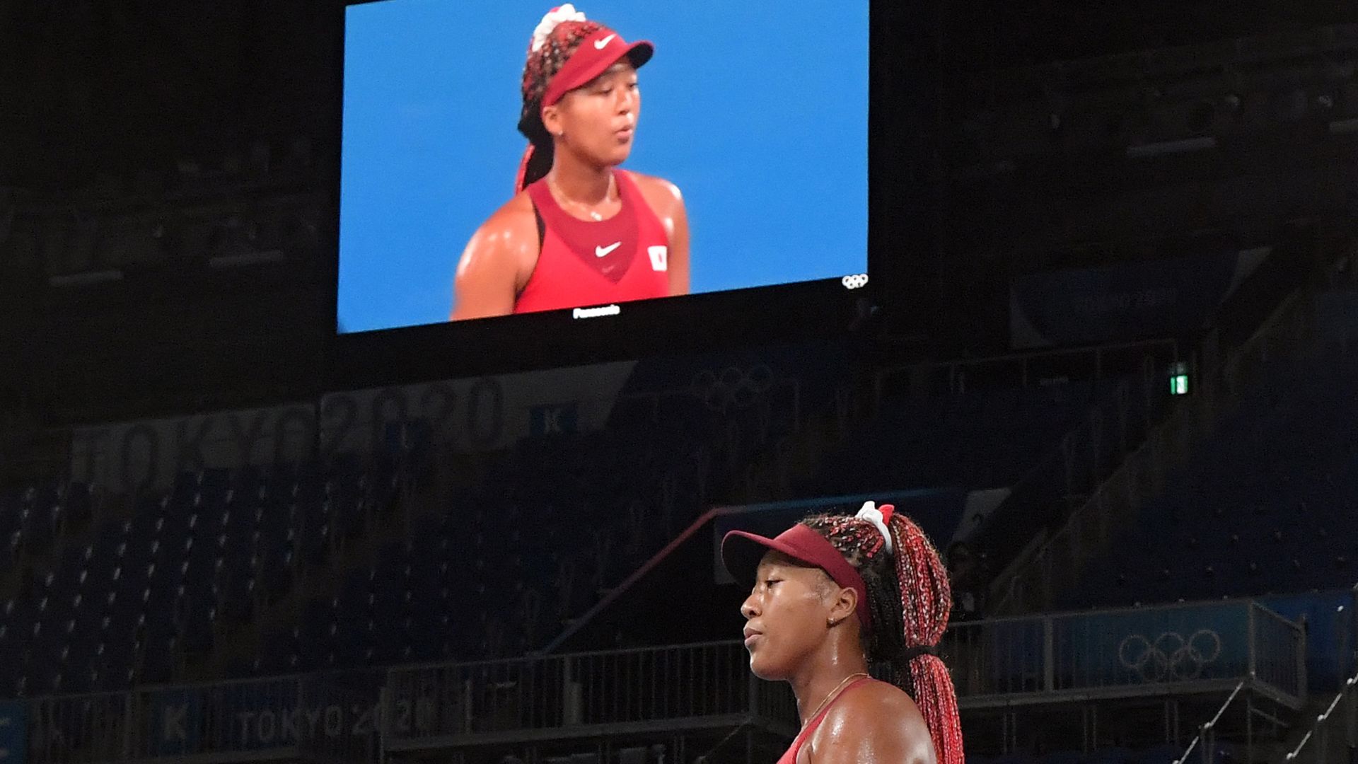 Photo of Naomi Osaka in her Olympic uniform breathing out a breath with a large screen reflecting her image