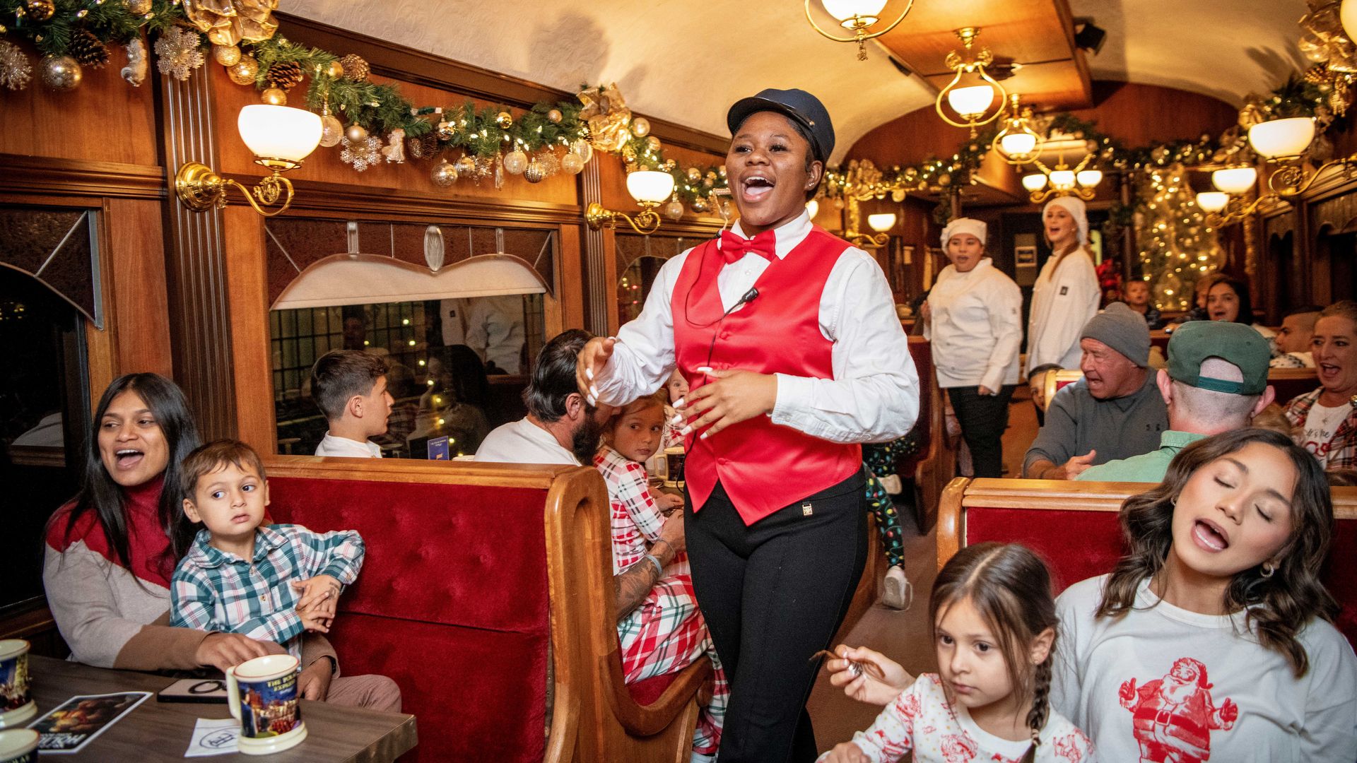 A festive train car decorated with Christmas garlands and lights. A woman in a red vest and bowtie sings to seated families, including children and adults, enjoying the holiday atmosphere.
