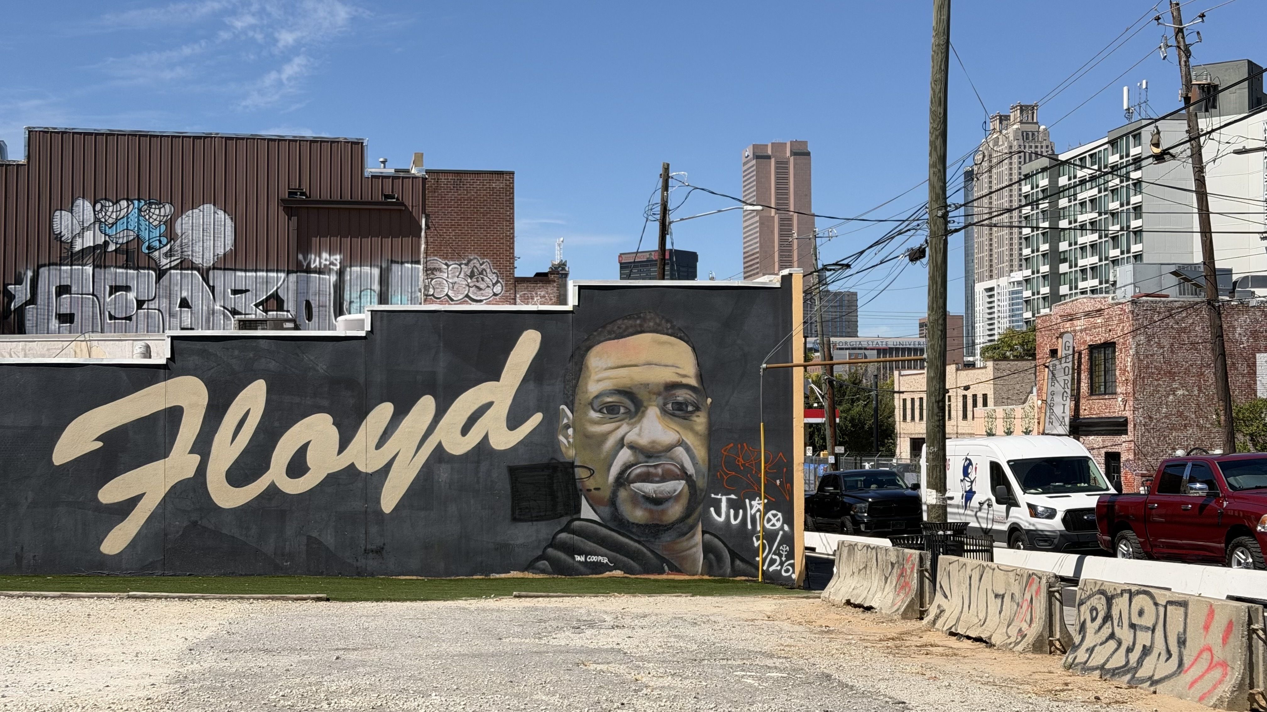 Urban mural with a large portrait and the word "Floyd" across a dark wall, graffiti on brick and metal panels, power lines, tall buildings in the distance, and parked vans and trucks along a street.