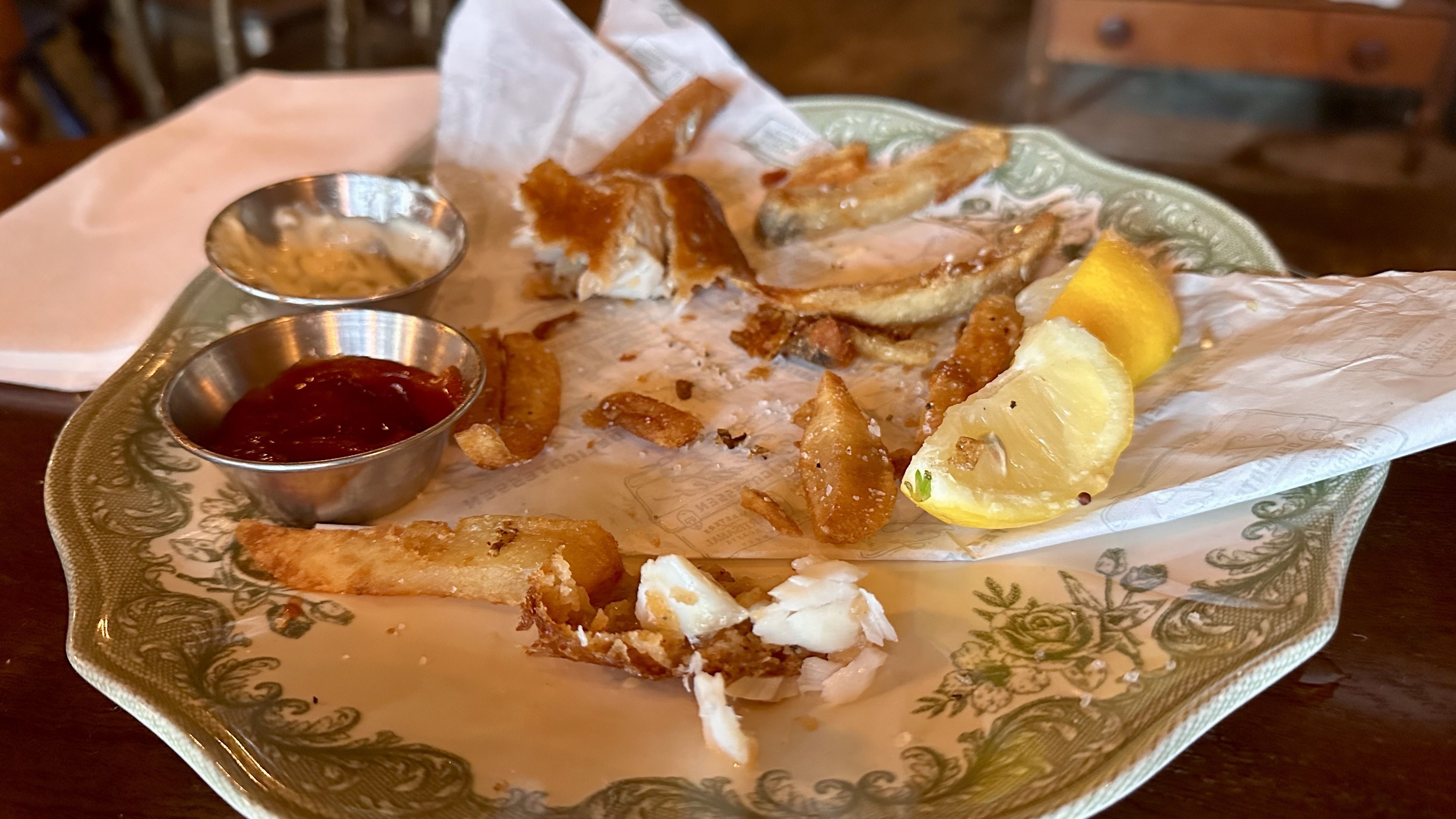 A nearly empty plate with leftover fries, fish pieces, a lemon wedge, a small container of ketchup, and another container with a creamy sauce on a wooden table.