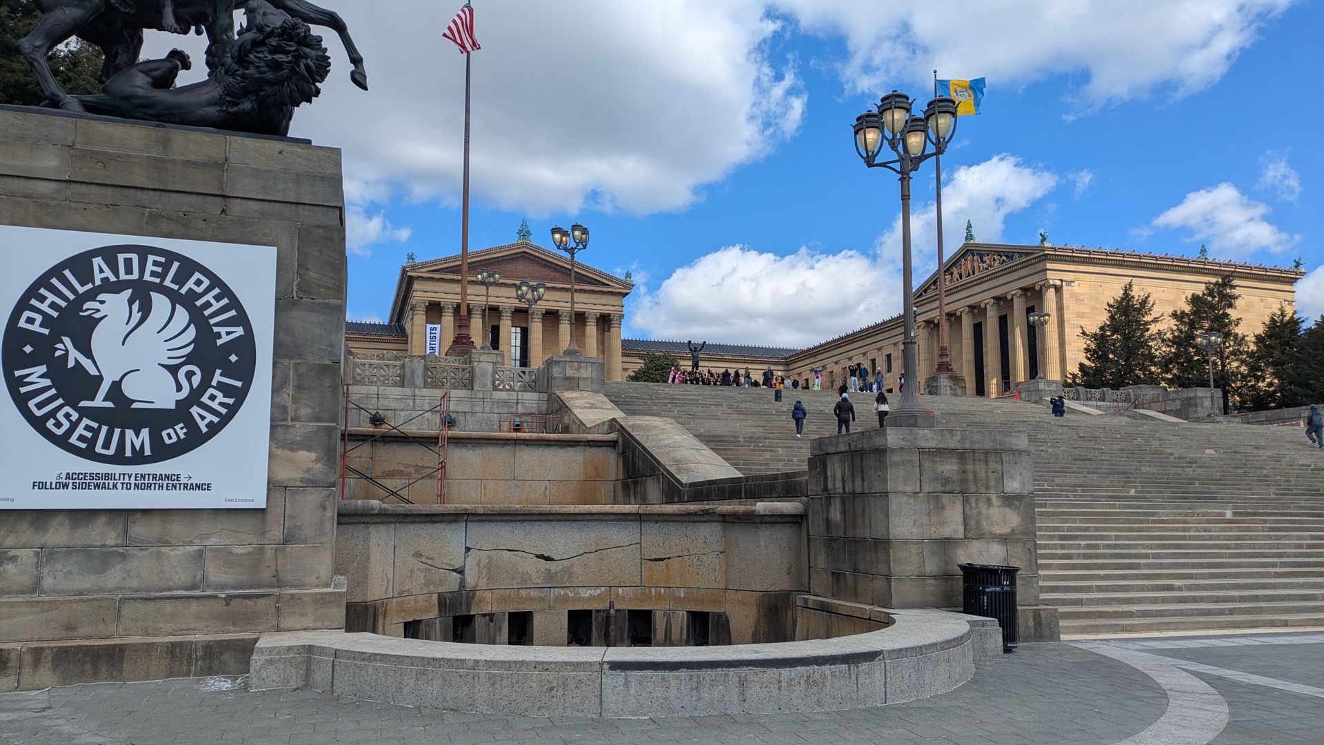 Wide view of the Philadelphia Museum of Art stairs and neoclassical buildings under a blue sky. A large circular PMA sign with a winged lion sits on a stone pedestal left; flags fly; people ascend the steps.