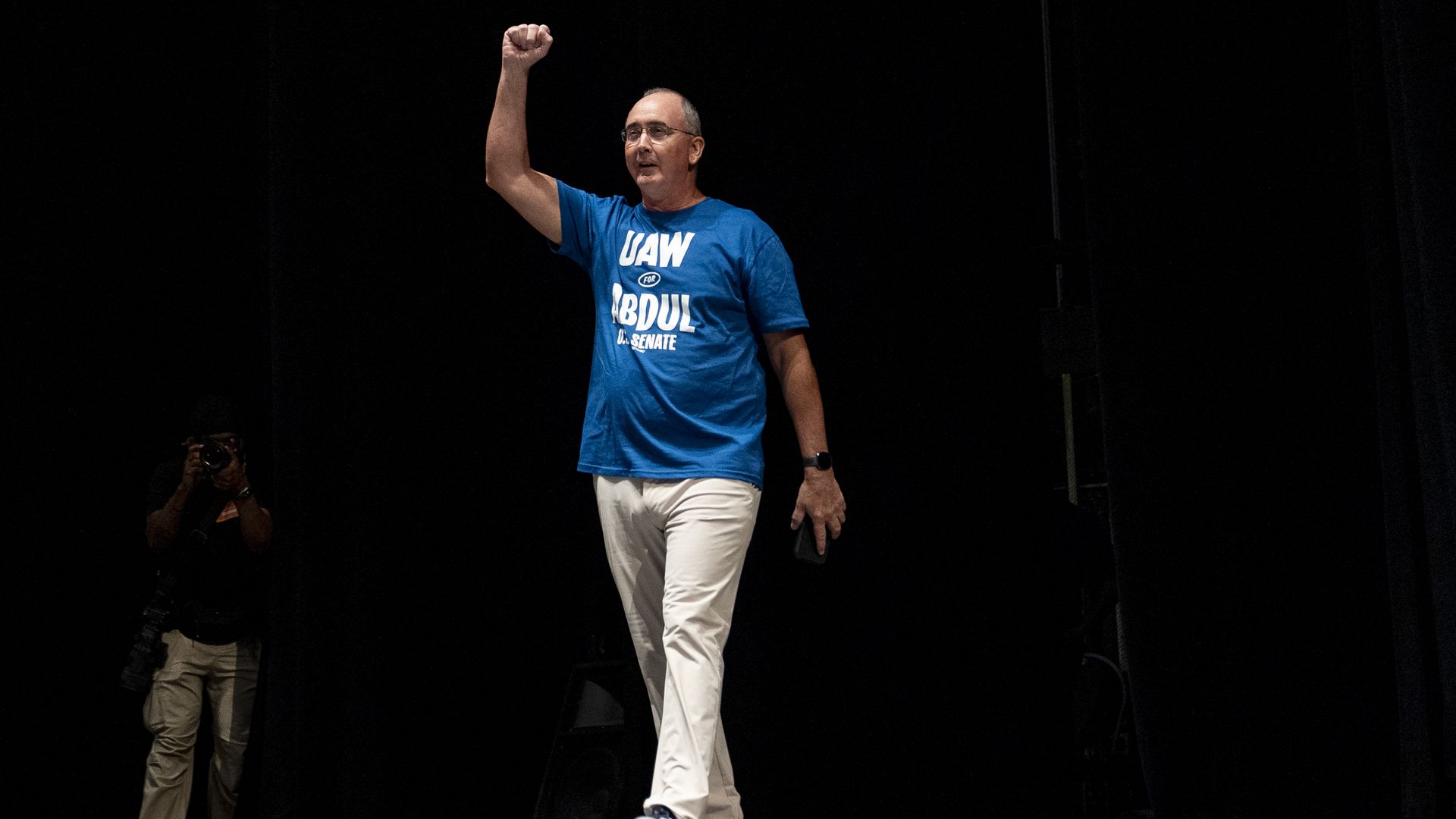 A person on a dark stage raises his right fist. He wears a blue T-shirt with white text, white pants, and sneakers; a photographer in beige pants shoots from the left foreground.