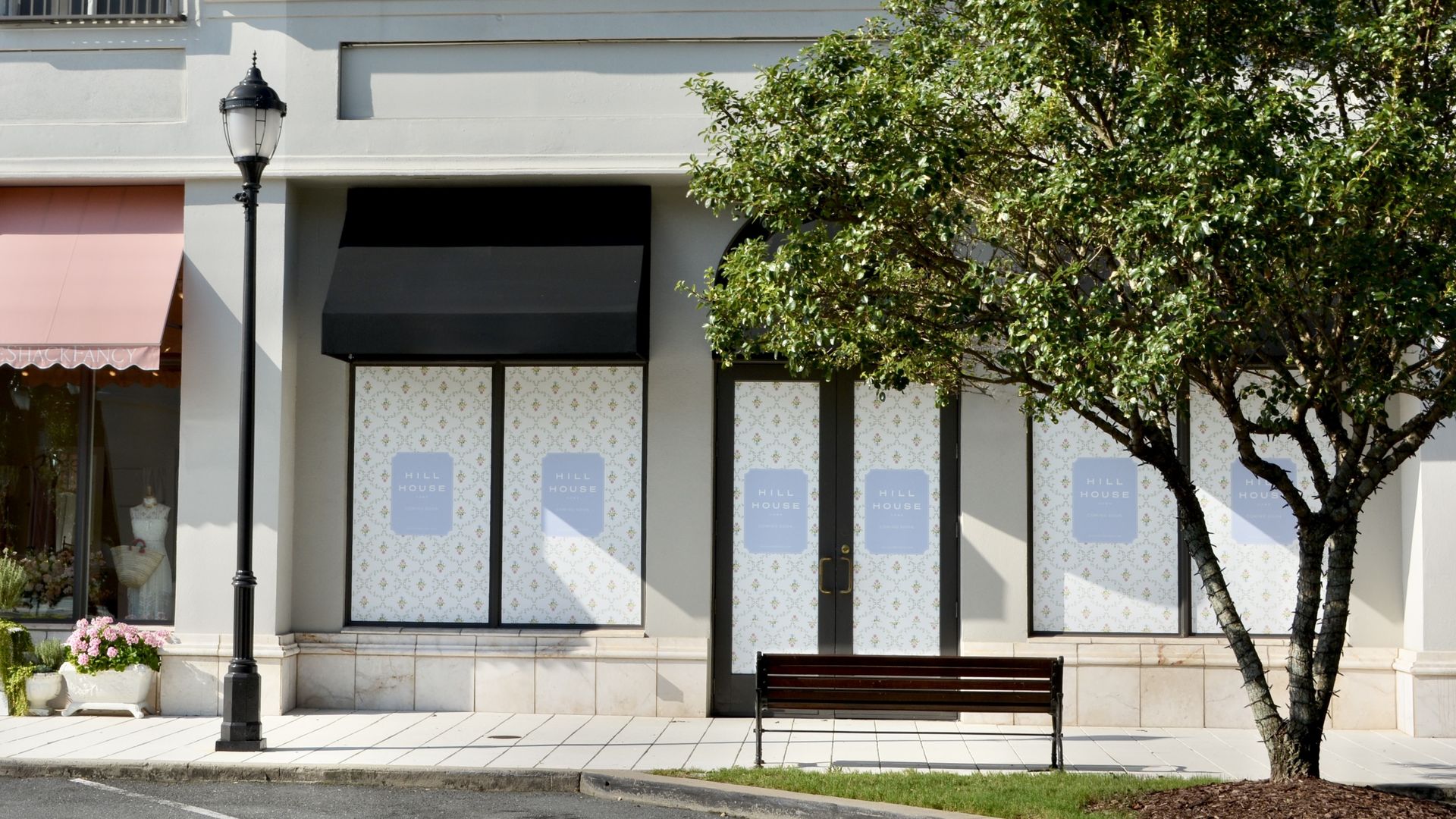 Empty storefront with closed doors and windows covered in patterned paper reading "HILL HOUSE," black awning above, a streetlamp and a bench near a leafy tree on a sunny day.