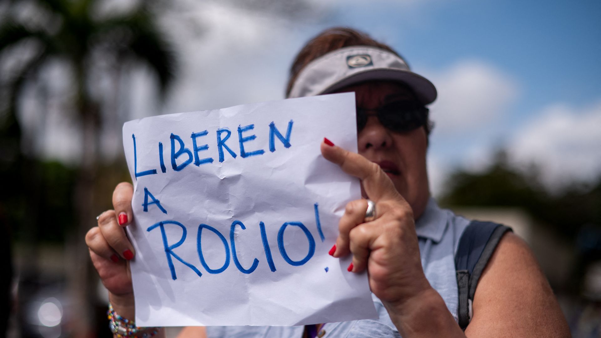 A woman in a sun visor holds up a paper sign that says "free Rocio" in Spanish. The paper is covering half of her face