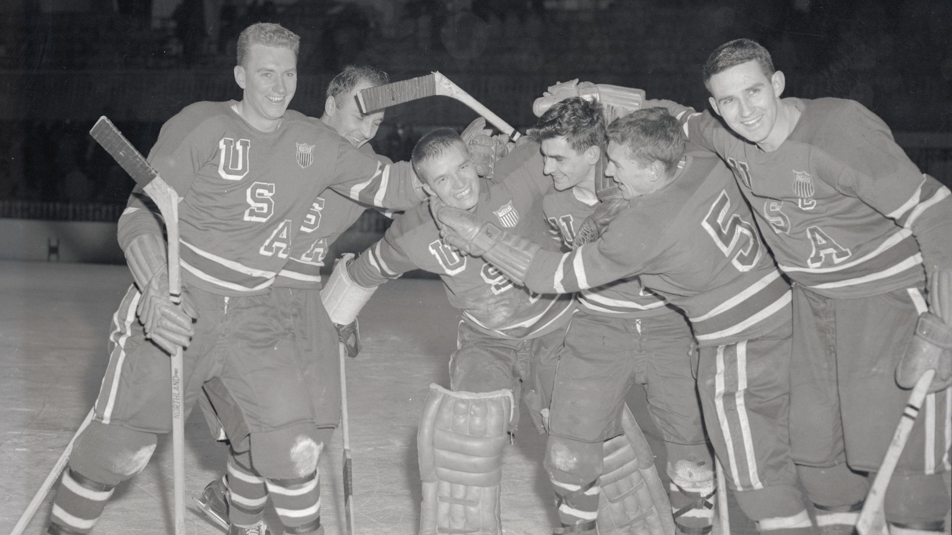 A black-and-white image of a hockey team in Team USA jerseys embracing a goaltender in 1950s-era leather hockey pads