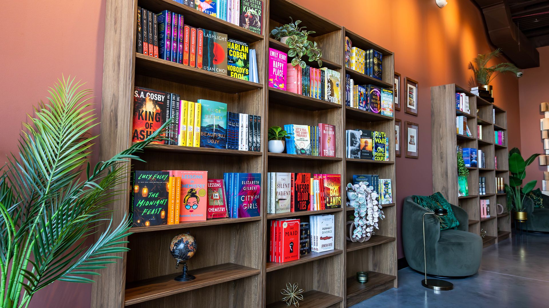 Warm bookstore interior with tall wooden shelves brimming with colorful book covers, orange walls, green plants, a small globe, and gray lounge chairs along the aisle.