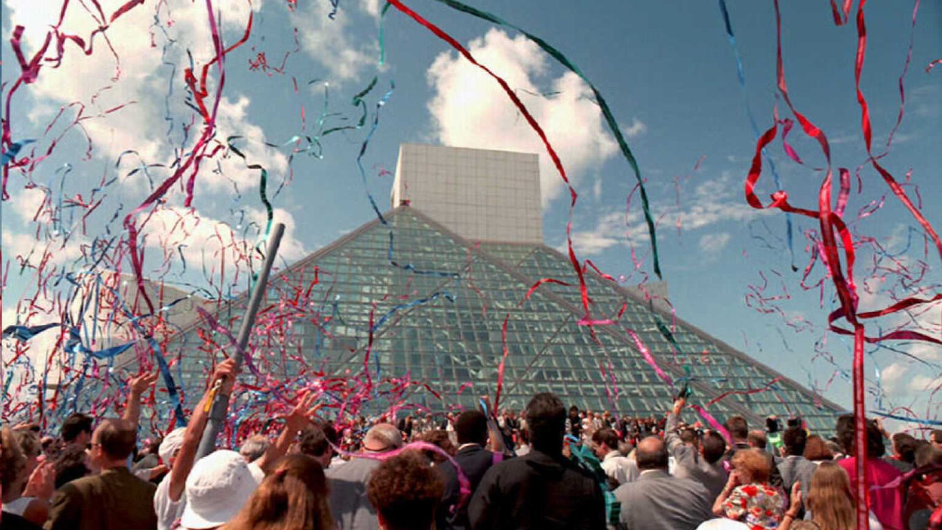 A crowd celebrates in front of a glass pyramid building with colorful red, blue, and pink streamers flying in the air under a partly cloudy blue sky.