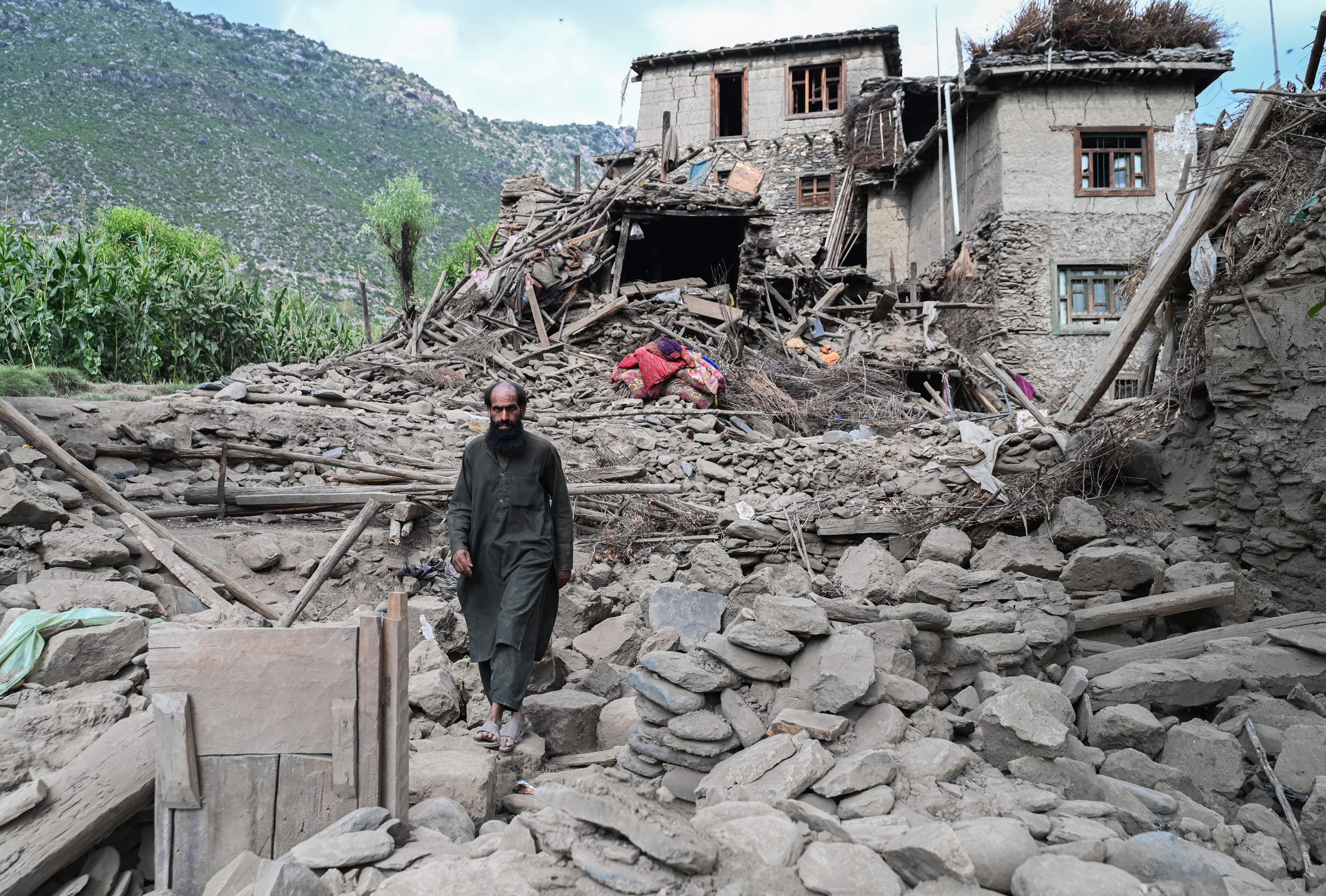 A man in traditional Afghan dress walks past a damaged house that's been reduced to rubble by an earthquake. Some houses are still standing to the right of it and a hill and vegetation can be seen behind it.