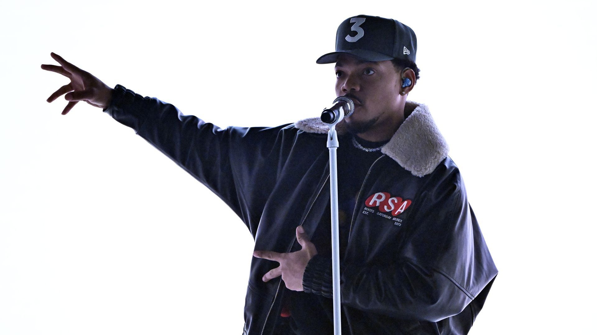 A photo of Chance the Rapper wearing a baseball hat with the number 3 on it and an oversized leather jacket singing into a microphone with his right arm raised and fingers spread