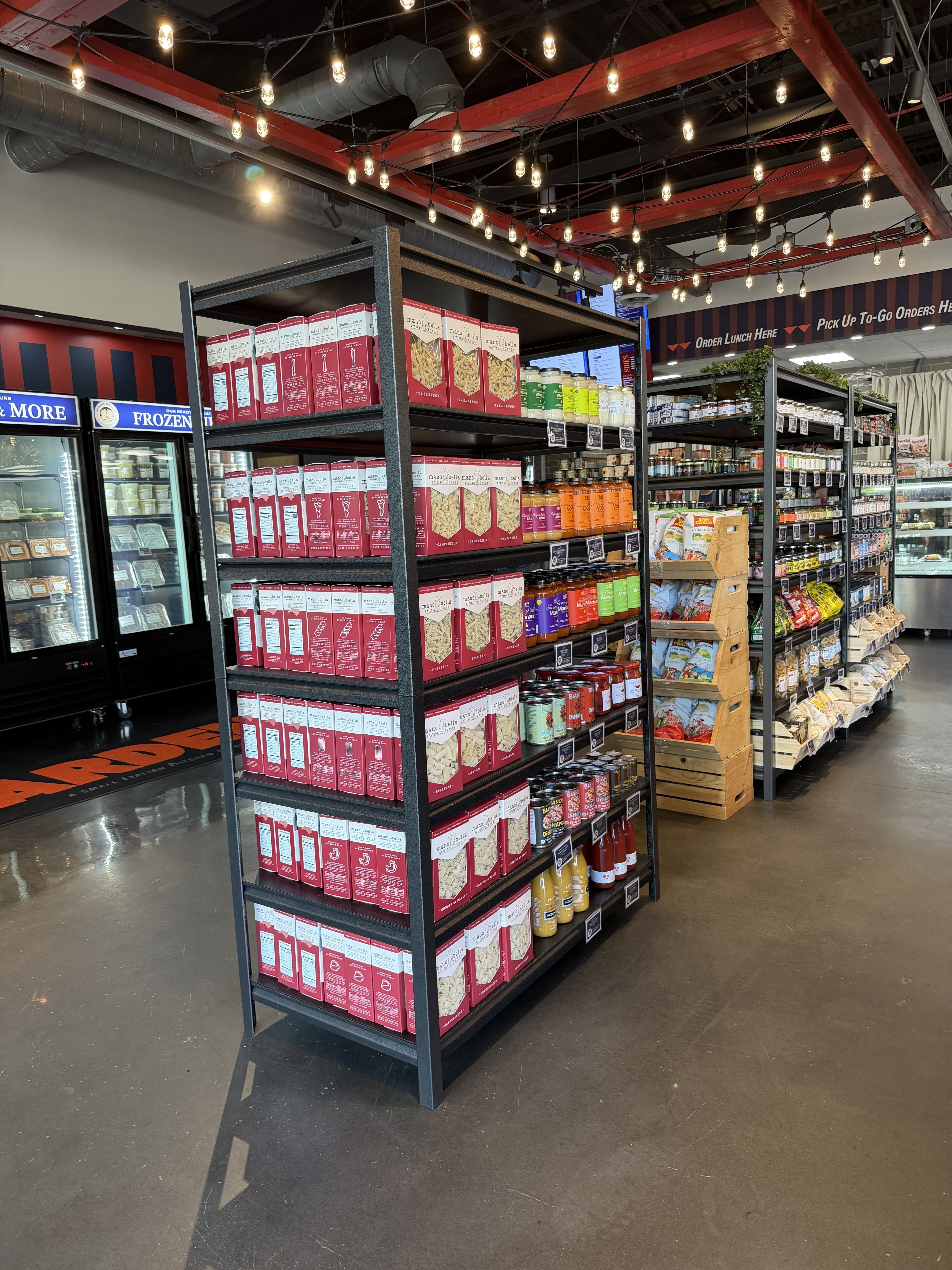 Interior of a grocery store with black metal shelves stacked with pink boxes, jars, and bottles. Warm string lights hang from red beams above, and a frozen display is to the left.