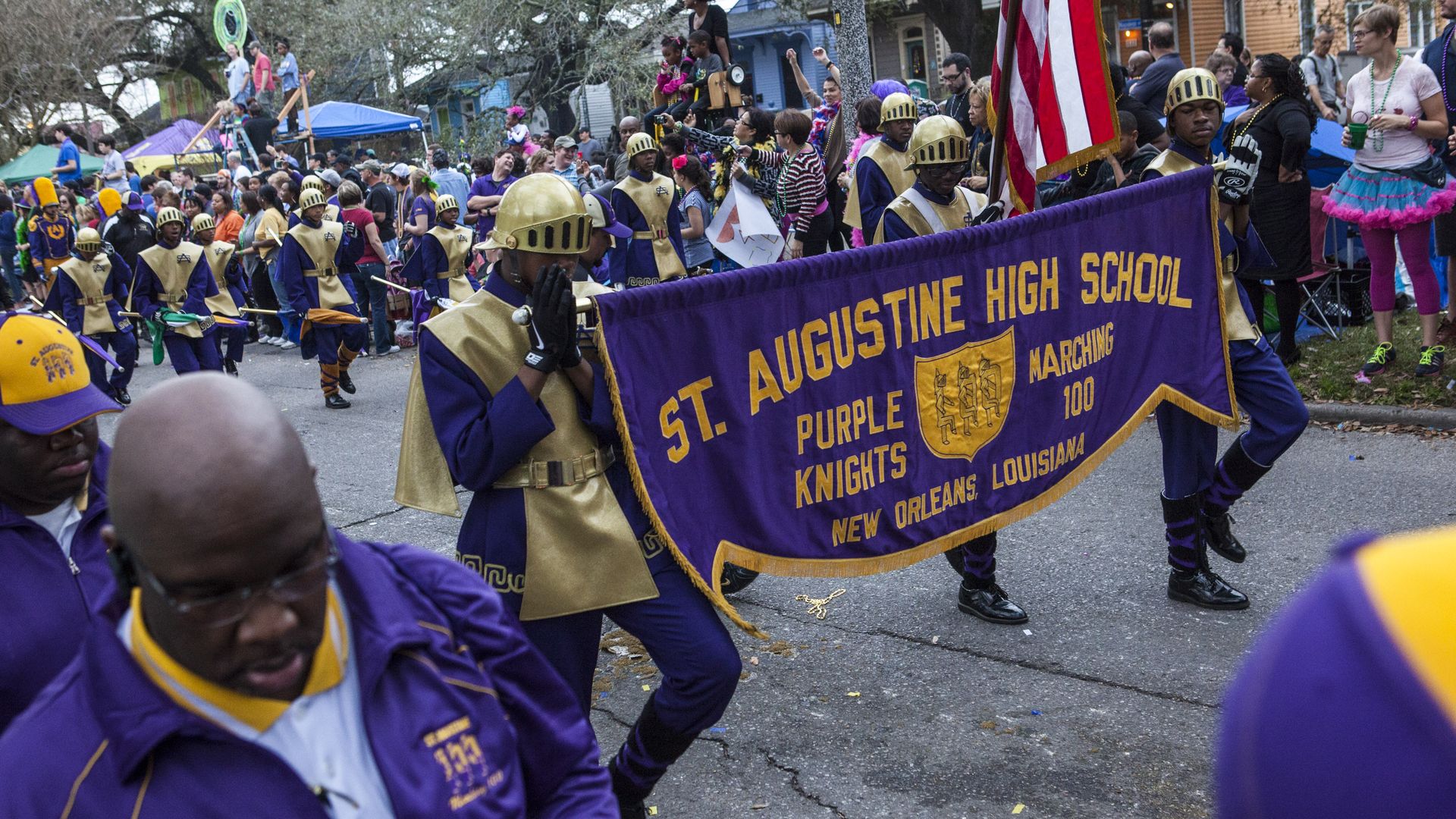 St. Augustine High School marching band students march in a Mardi Gras parade. Two of them hold a banner with the school name at the front of the band.