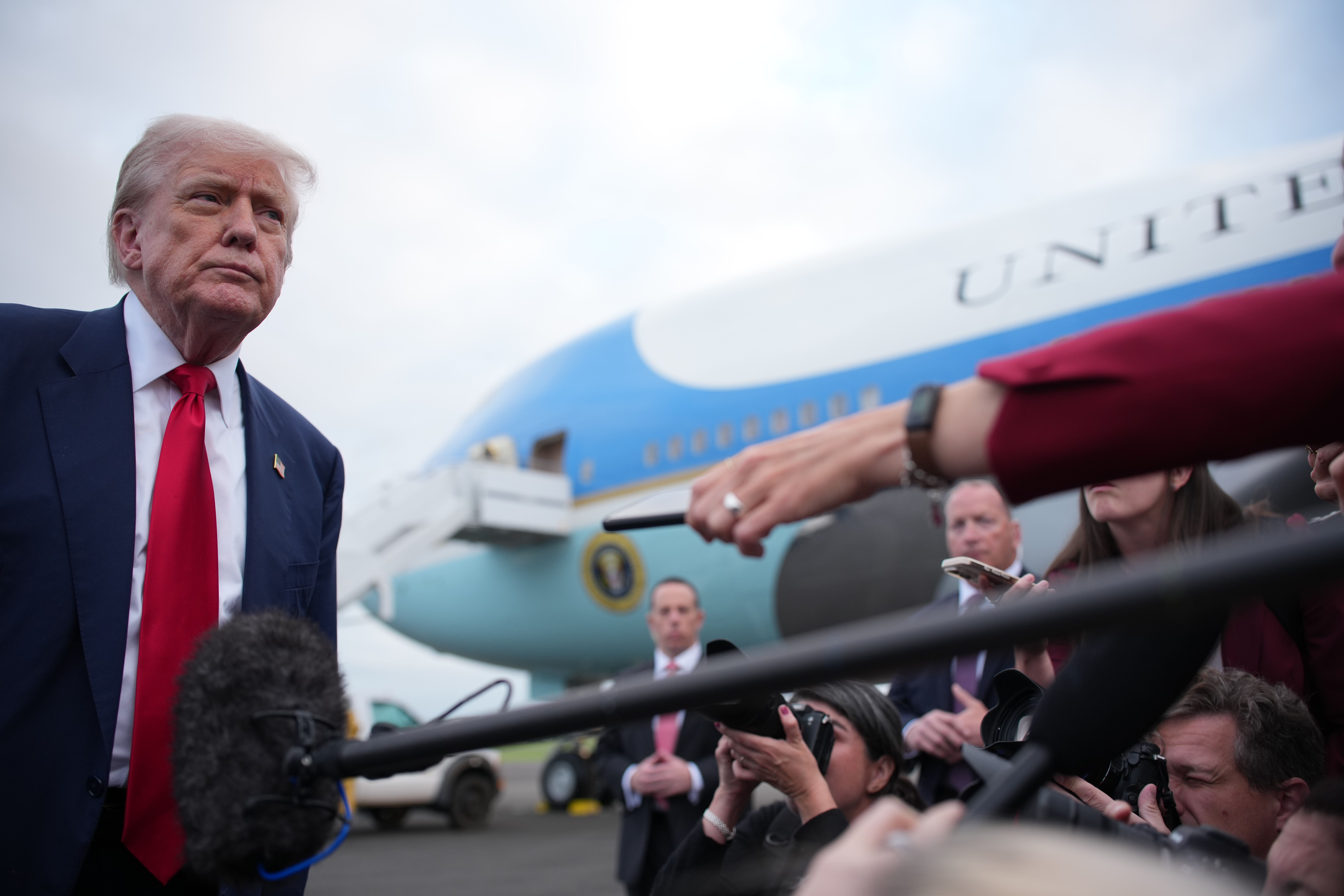Former President Donald Trump in a dark suit and red tie speaking to reporters with microphones and cameras, with Air Force One plane in the background on cloudy day.