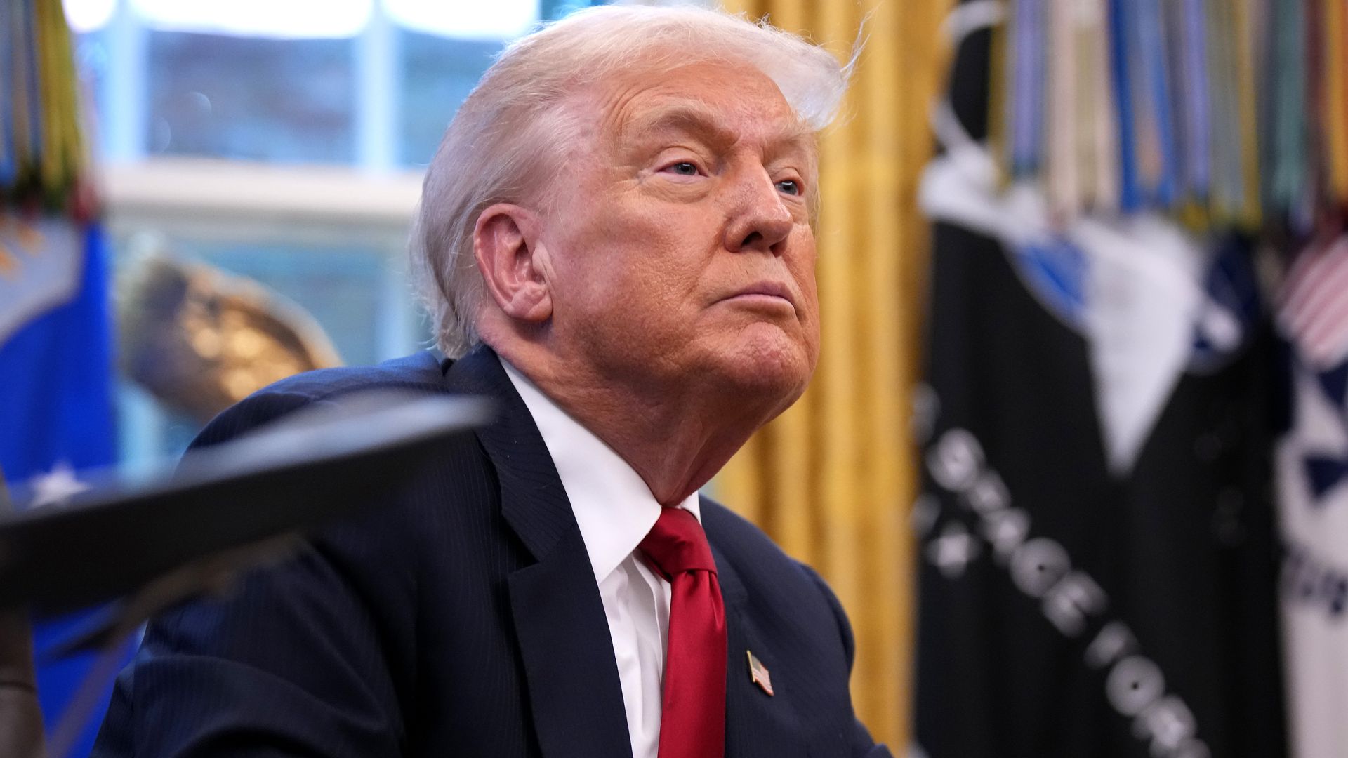 Donald Trump in a dark suit, white shirt, and red tie sitting at a desk with hands clasped, looking upward in a room with gold curtains and flags in the background.