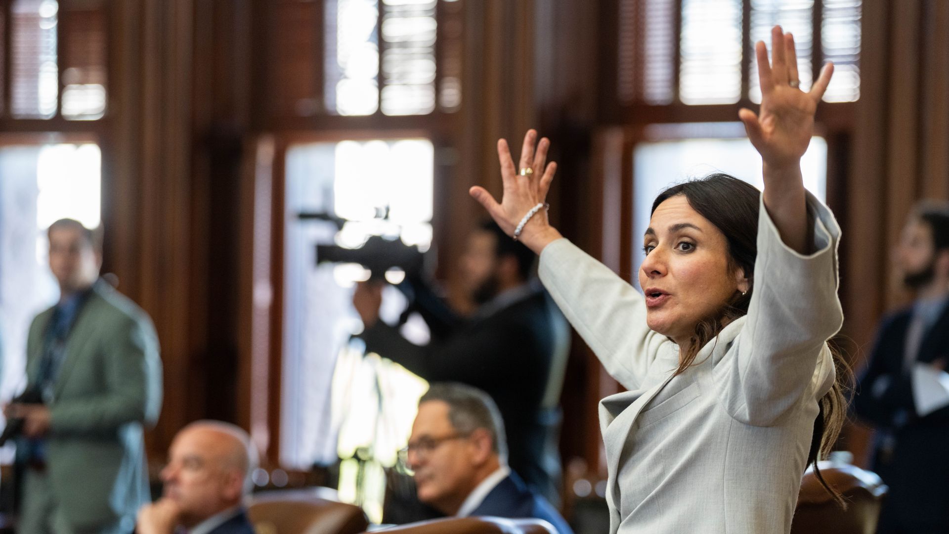 A woman wearing a white suit puts her hands up in the Texas House Chamber.
