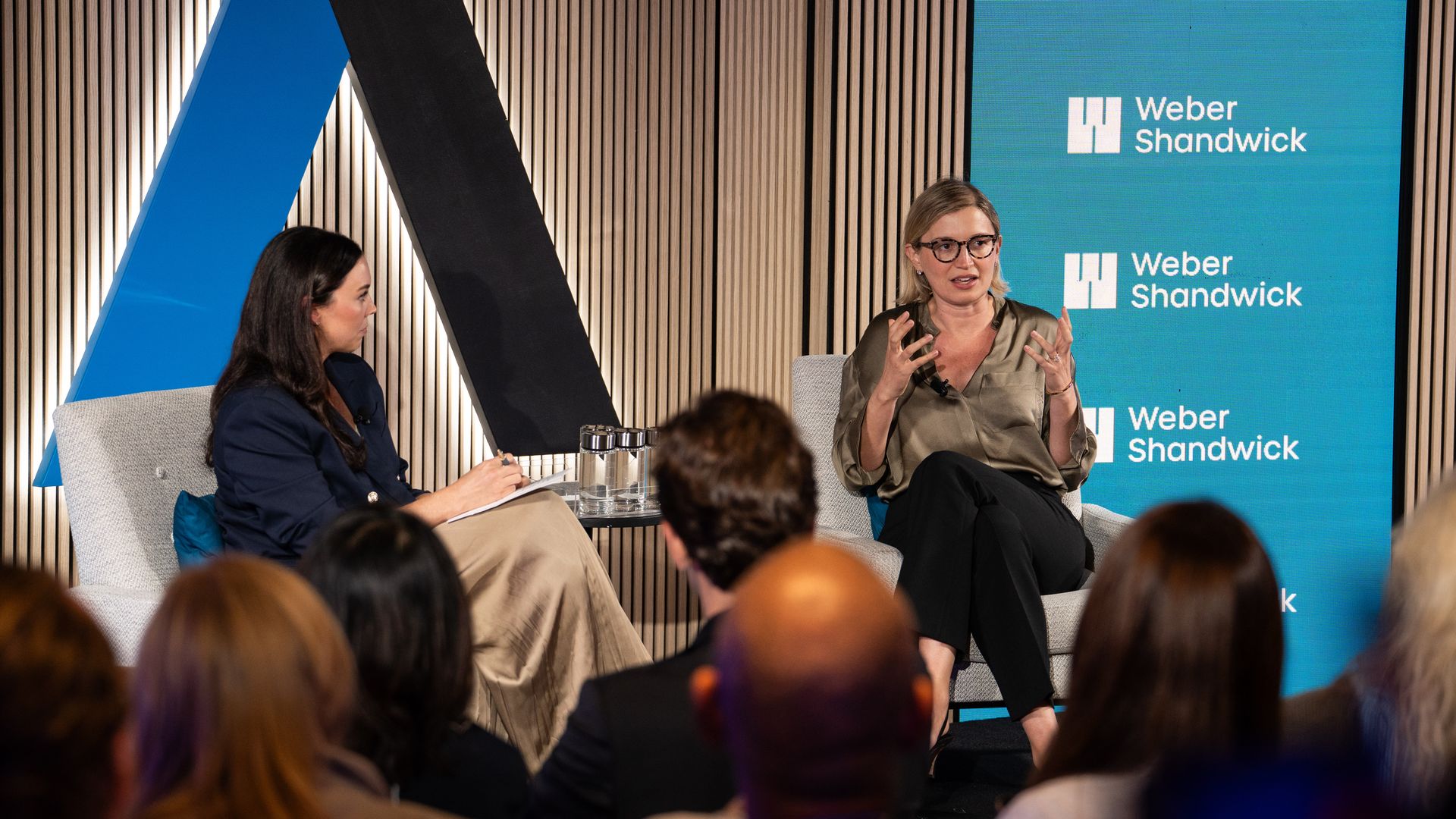 Two women in conversation on a stage; one wears a dark blazer and skirt, the other wears glasses, a light olive blouse, and black pants, with a large blue and black A-shaped structure and a "Weber Shandwick" screen behind them.