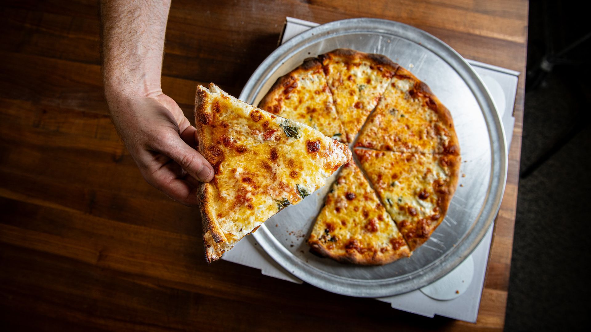 A hand holds a piece of pizza above a whole pie that's on a metal plate on a table
