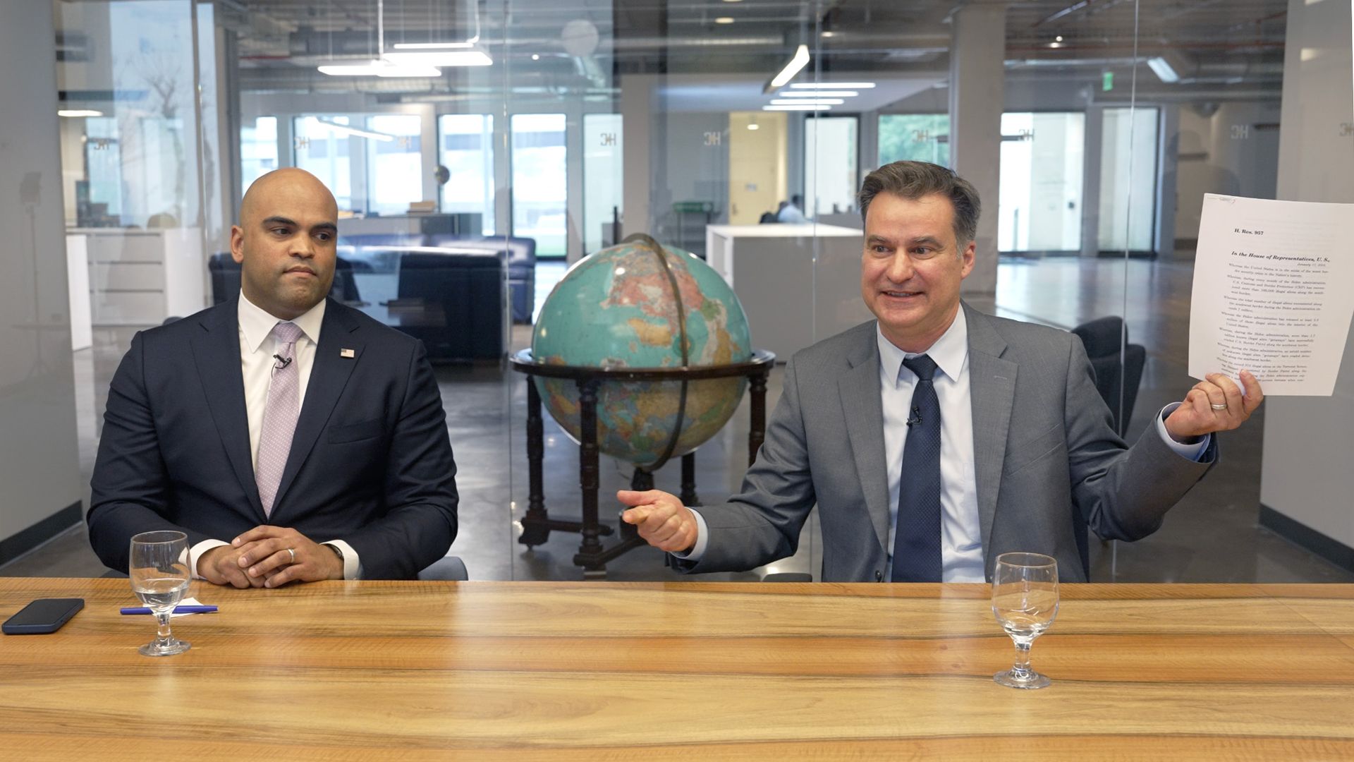 Two men sit at a conference table. One holds up a piece of paper.