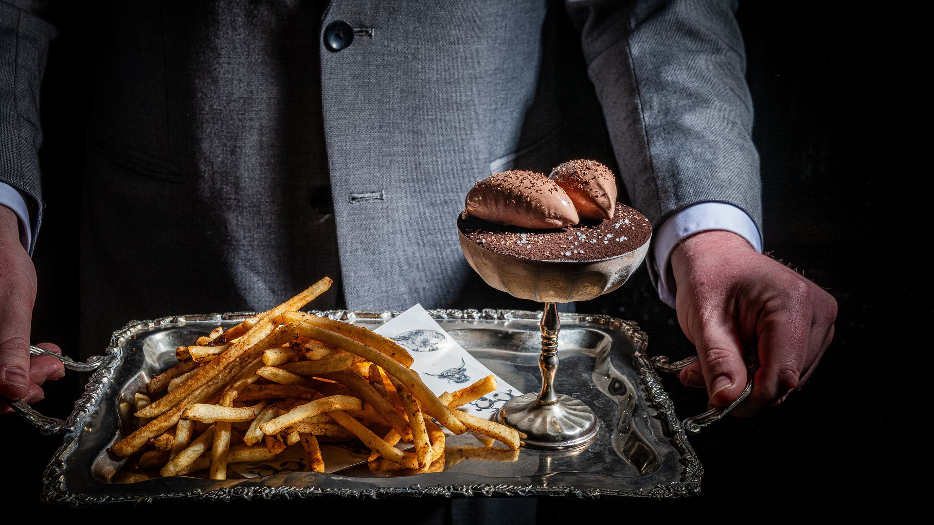 Person in a gray suit presents a silver tray: left piled French fries, right a chocolate dessert in a coupe glass with cocoa dust and two small toppings, set against a dark background.