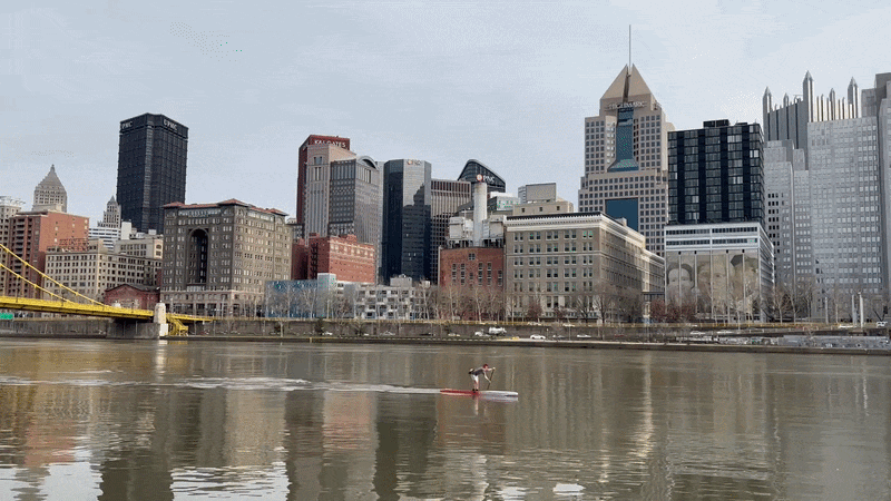 A man on paddling on a stand up paddle board in a river with an urban skyline and yellow bridge in background.