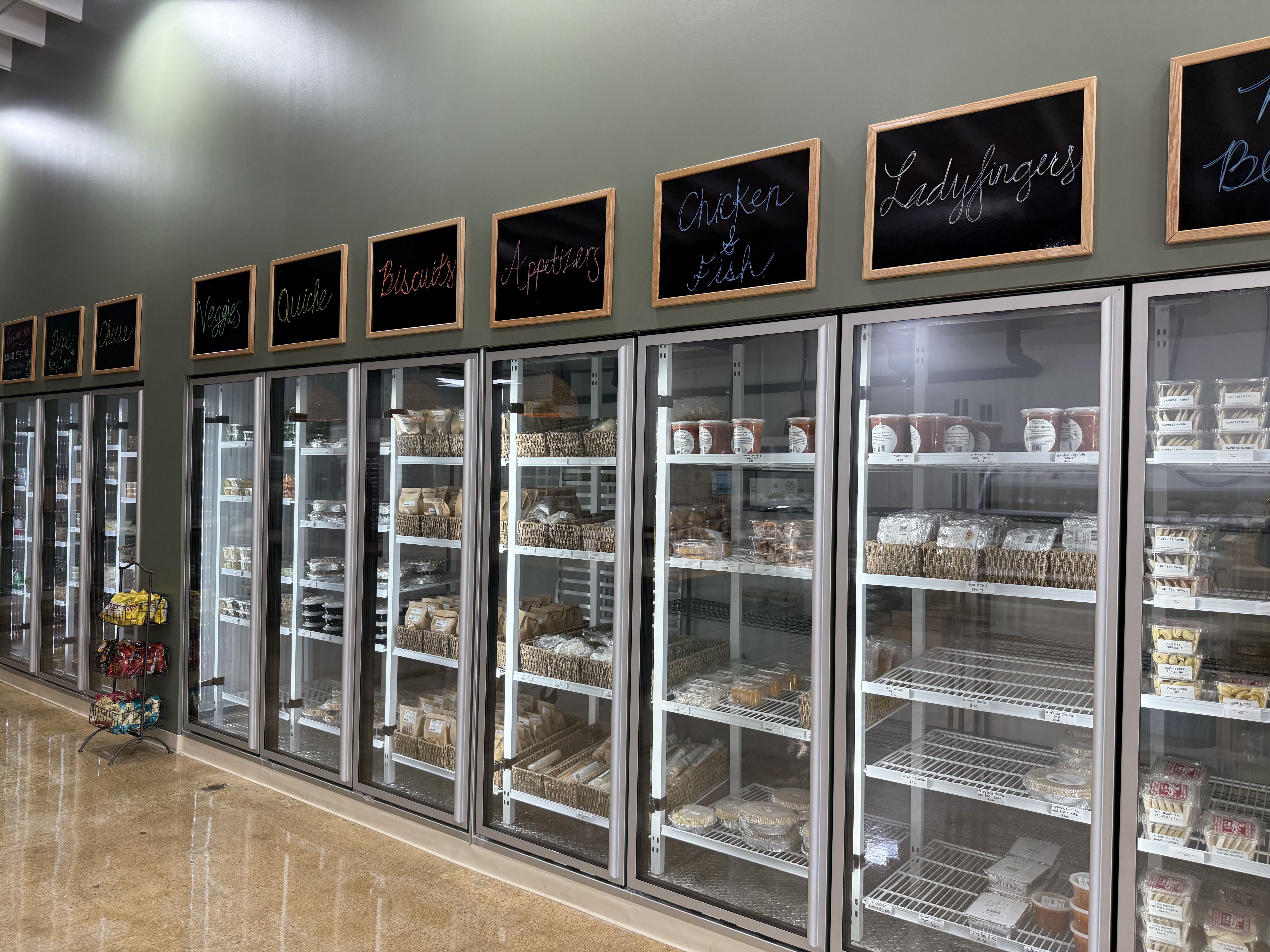 A row of glass-door refrigerated shelves in a store with sections labeled Veggies, Quiche, Biscuits, Appetizers, Chicken & Fish, and Ladyfingers, stocked with packaged food items.