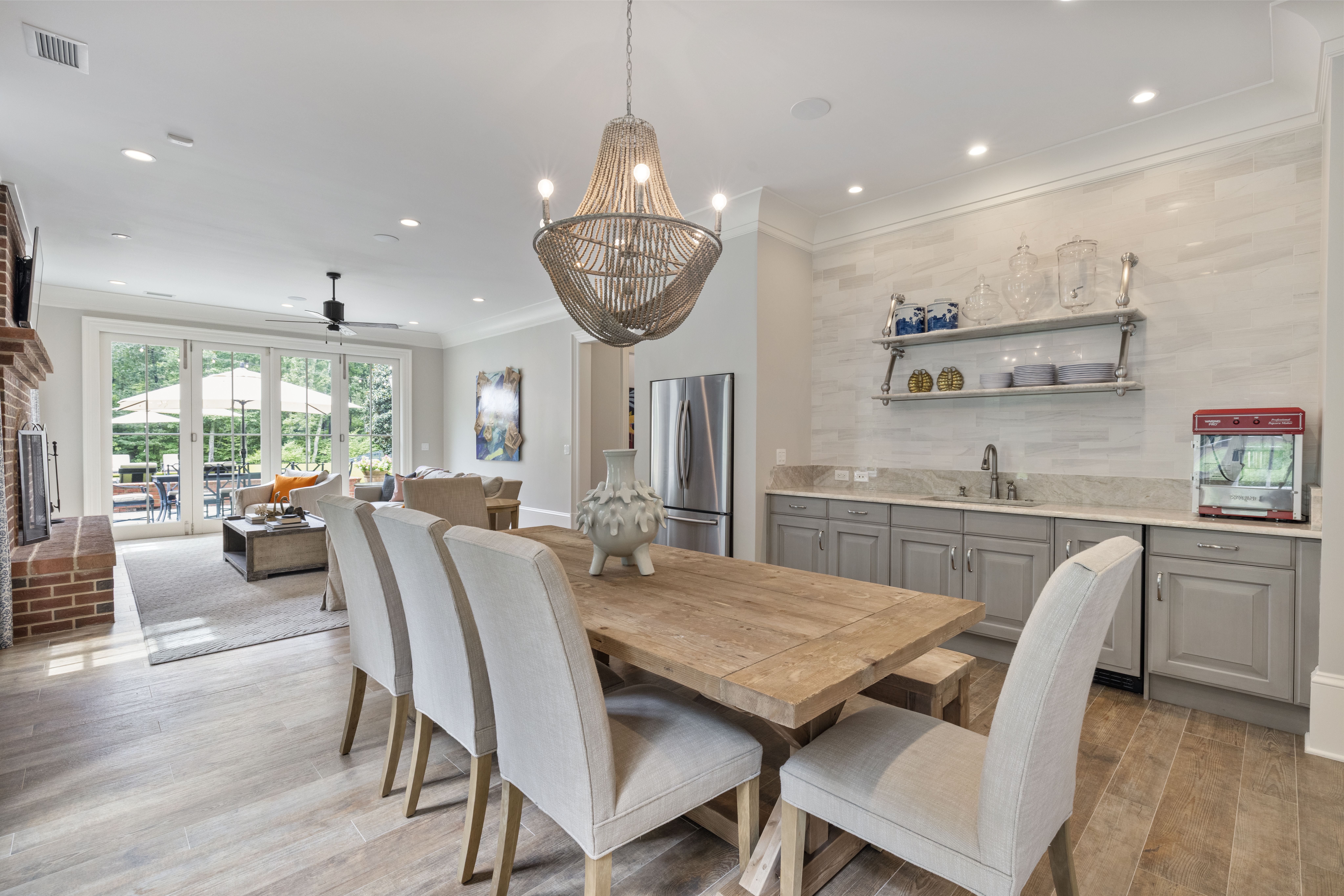 Bright open dining and living area with a wooden table, gray upholstered chairs, chandelier, gray cabinets, stainless fridge, white backsplash, and glass doors leading outside.