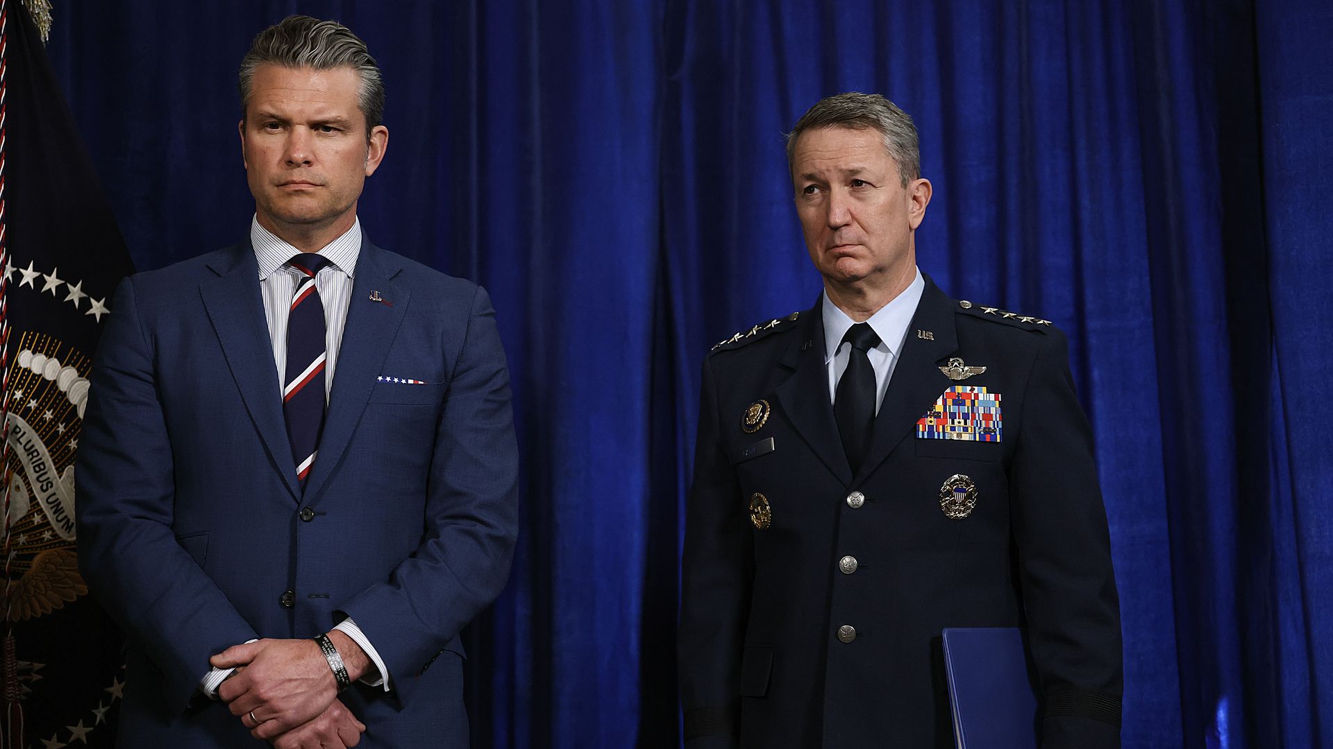 Two men stand before a blue curtain. One wears a blue suit with a striped tie, the other a decorated military uniform holding a blue folder beside a flag with a presidential seal.