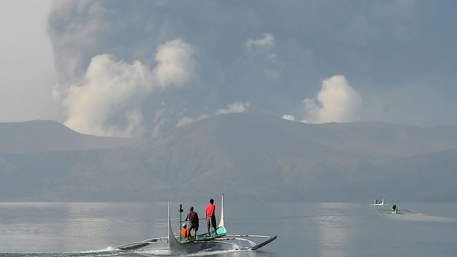 Residents living at the foot of Taal volcano ride outrigger canoes to reach their homes while the volcano spews ash as seen from Tanauan town in Batangas province, south of Manila