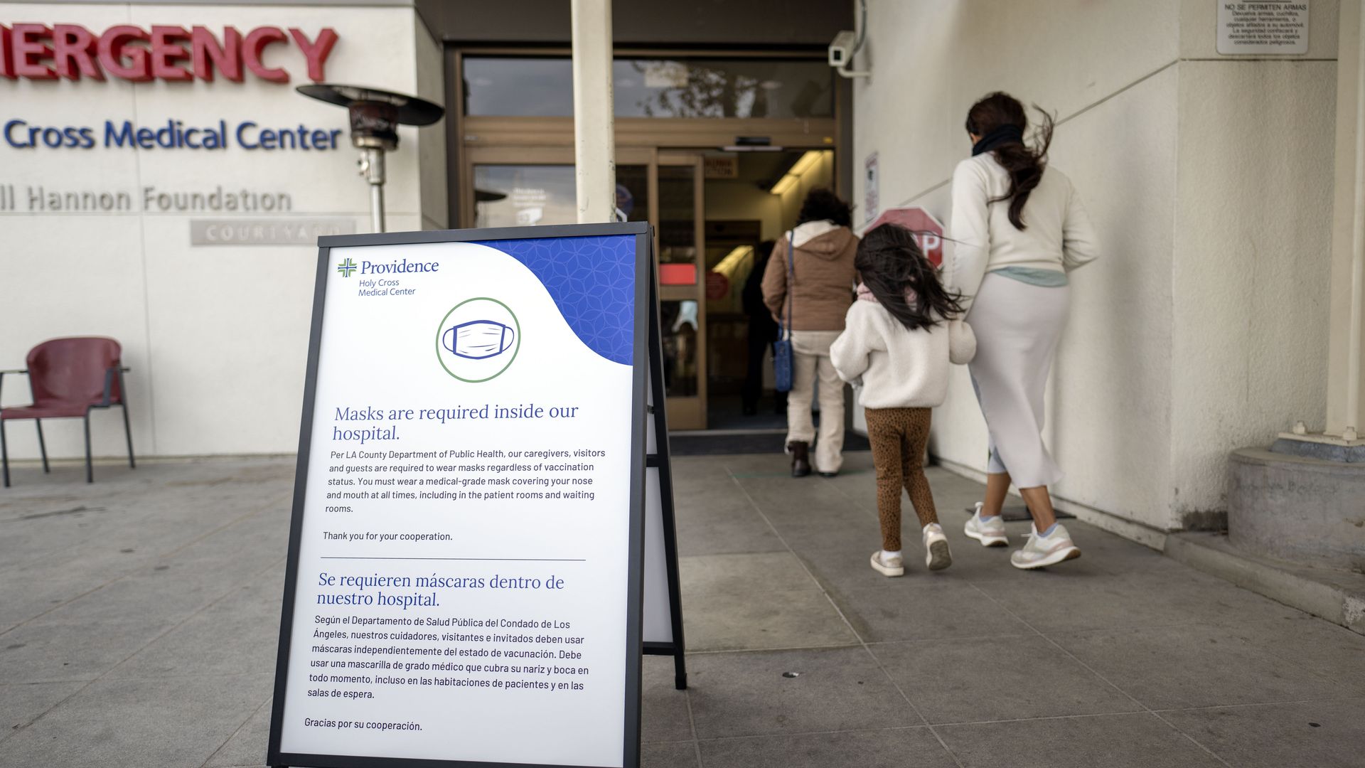 A woman and child walk on the right side of the photo into a hospital door. A sign outside the door posted on the floor says masks are required. On the left, the word "emergency" in red. 