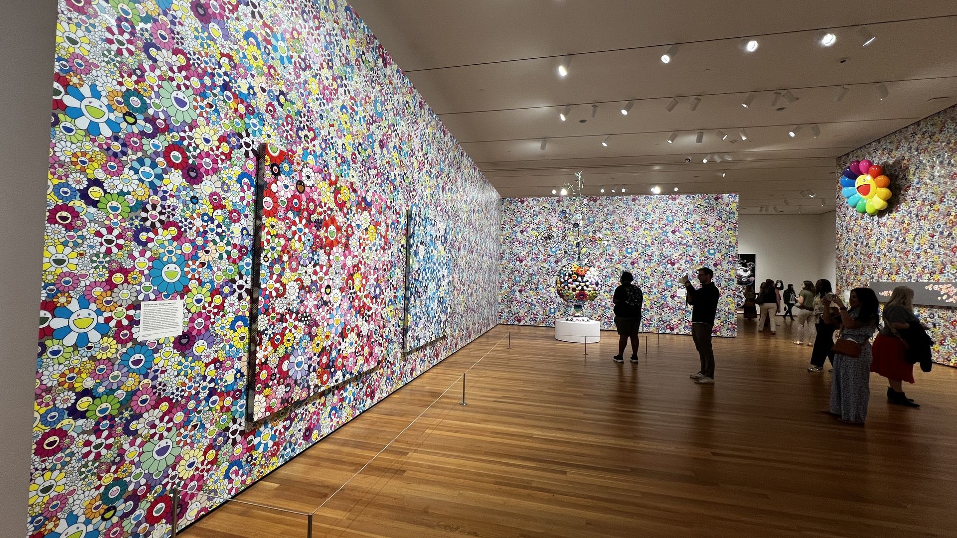 Gallery interior with walls covered in dense, multicolored flower patterns and smiling faces; visitors view large installations and a rainbow flower sculpture in a bright, wood-floored room.