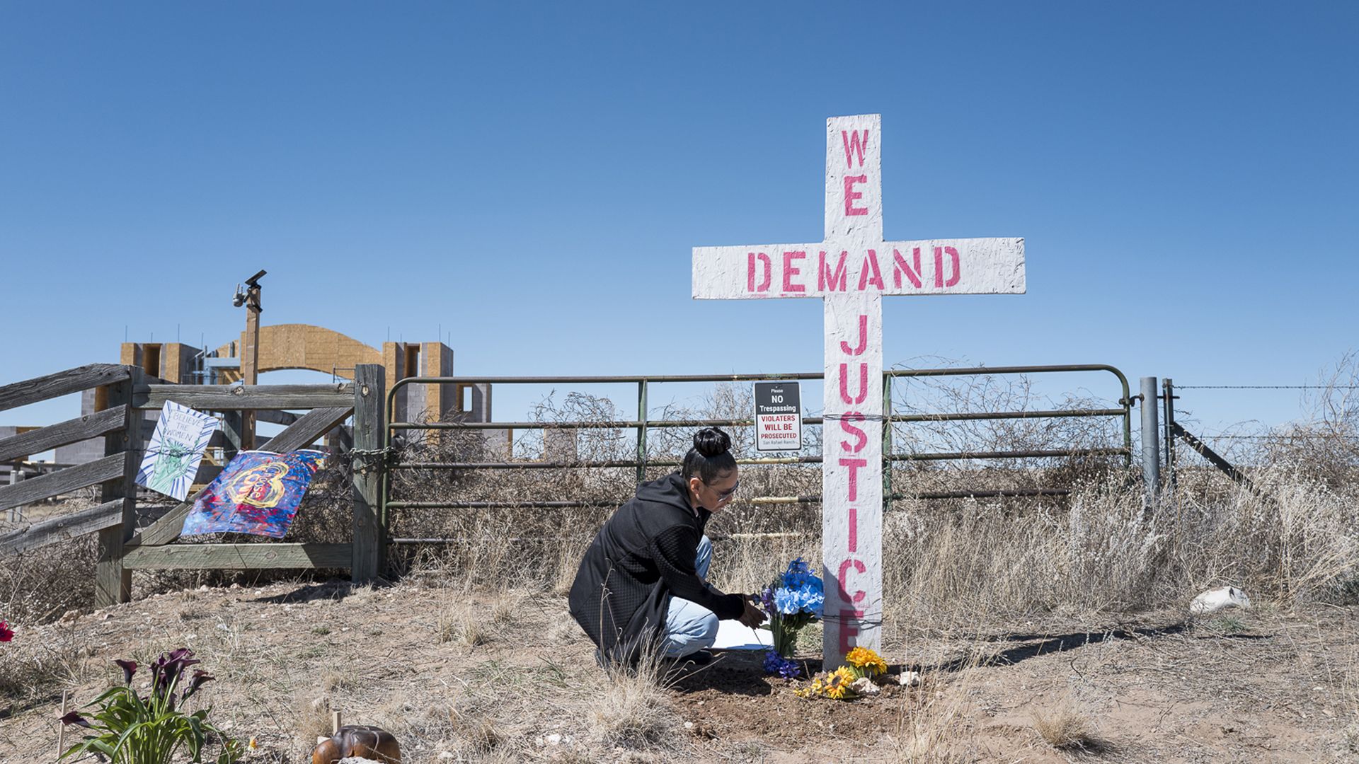 White cross-shaped sign with pink text reading WE DEMAND JUSTICE stands in a desert yard by a fence. A person kneels at its base placing flowers; a clear blue sky above.