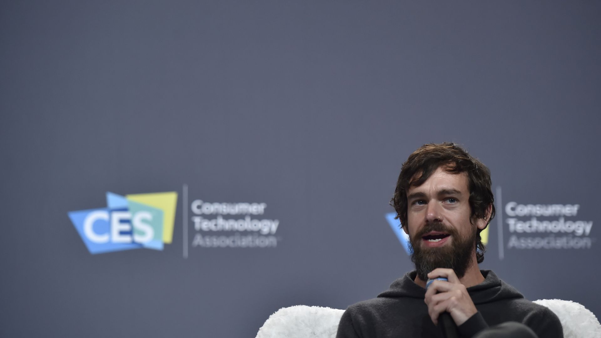 In this image, Twitter CEO Jack Dorsey sits in front of a dark gray wall at a conference, speaking into a microphone.