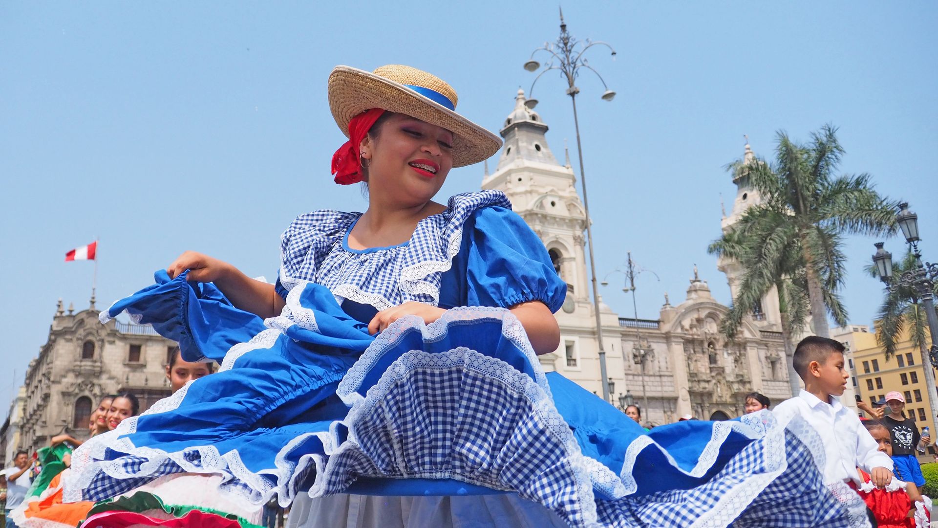 Smiling dancer in a blue-and-white gingham dress with lace, wearing a straw hat with blue ribbon and a red scarf, dancing in a sunny street parade near historic buildings and palm trees.