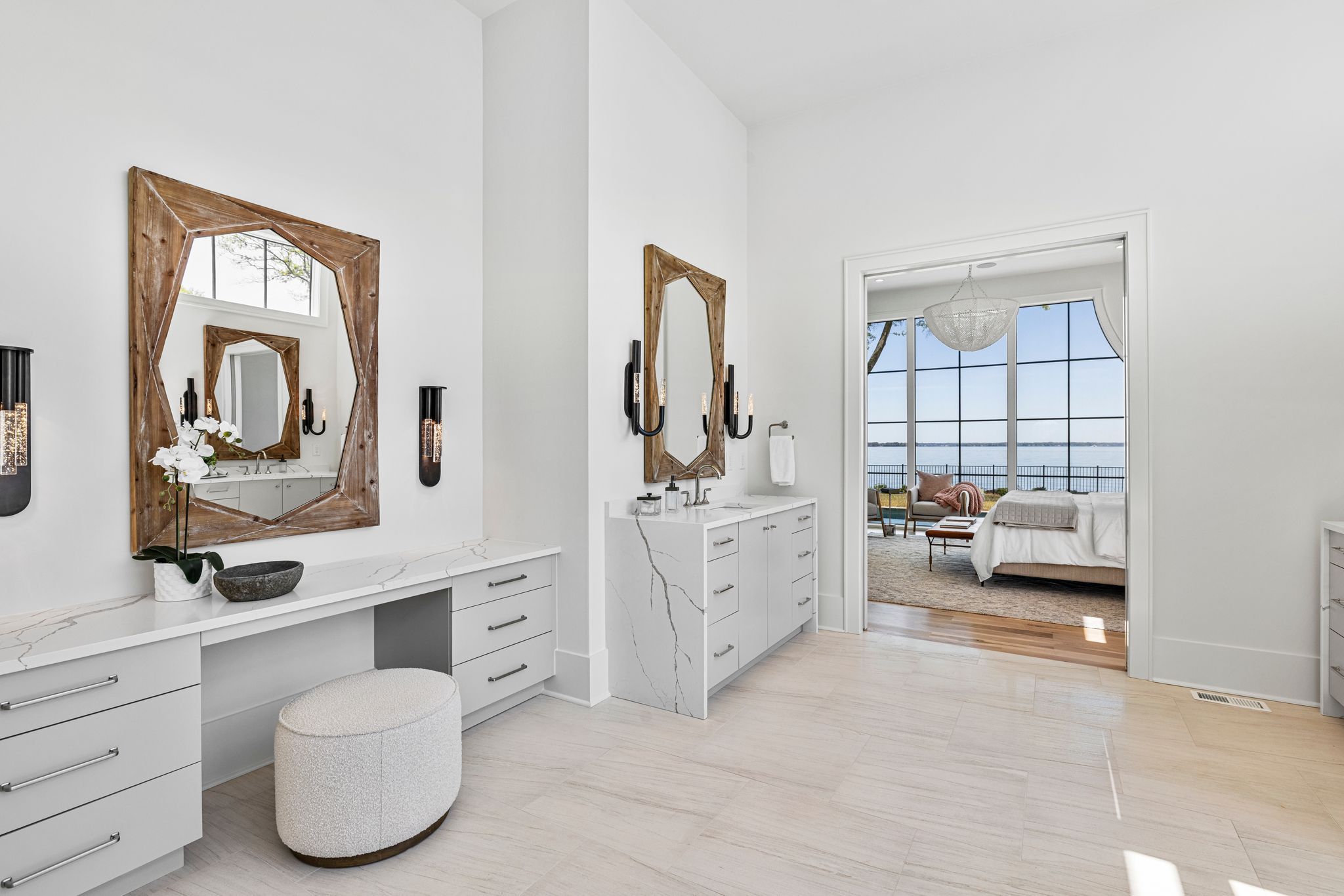 Bright modern white bathroom with marble countertops, wooden-framed mirrors, black wall sconces, and a view into a bedroom with large windows overlooking water.
