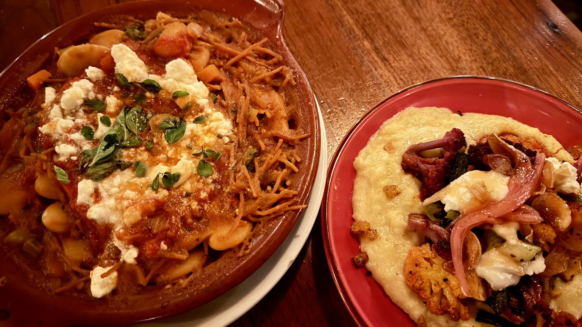 Two plates of warm food on a wooden table: a large oval baking dish filled with pasta in tomato sauce, ricotta, and herbs; a red plate with cheesy flatbread topped with ham and onions.