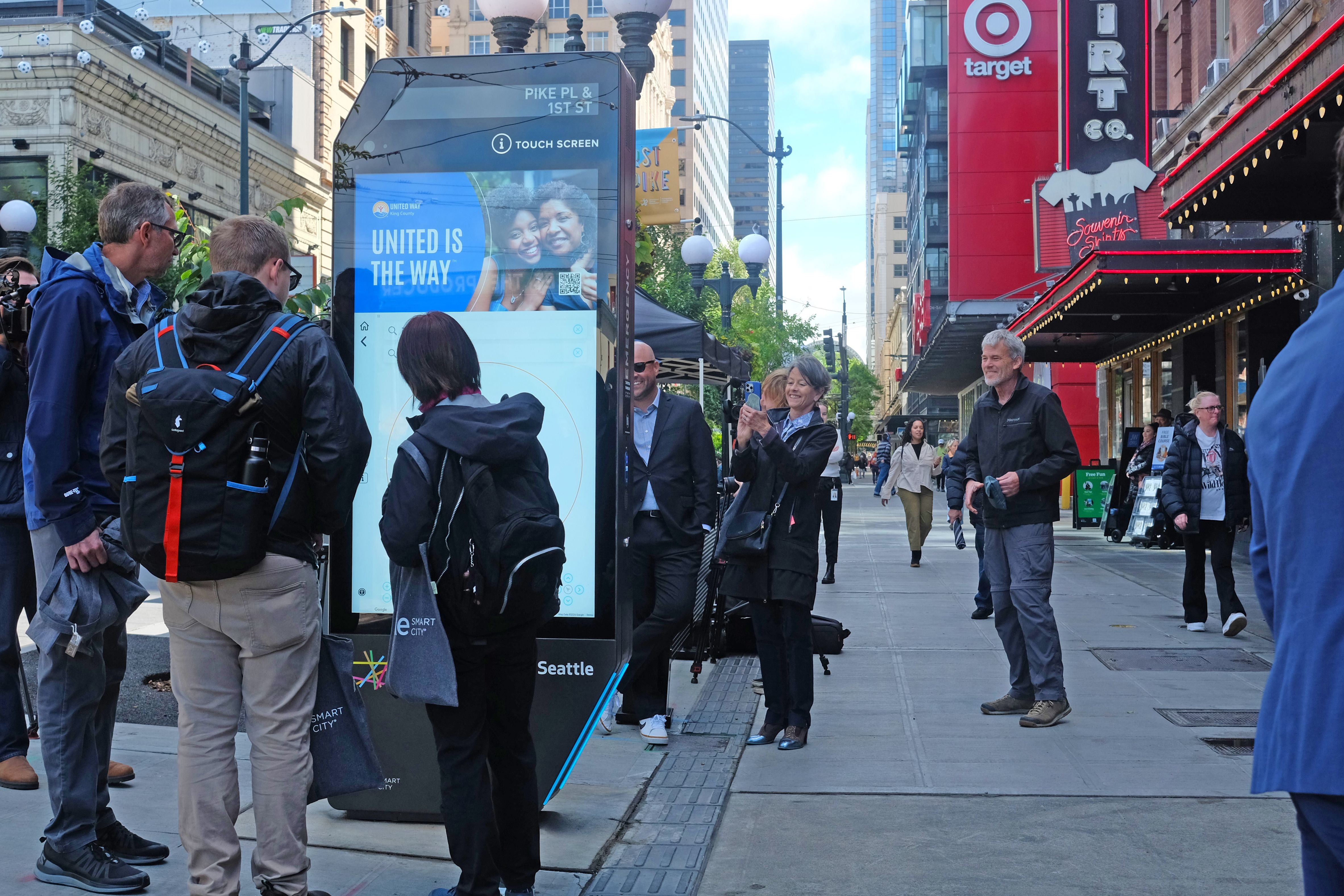 Busy city street with a large interactive kiosk in the center showing "UNITED IS THE WAY." People gather, smile, and take photos. A red theater front with string lights and a Target sign on the right.
