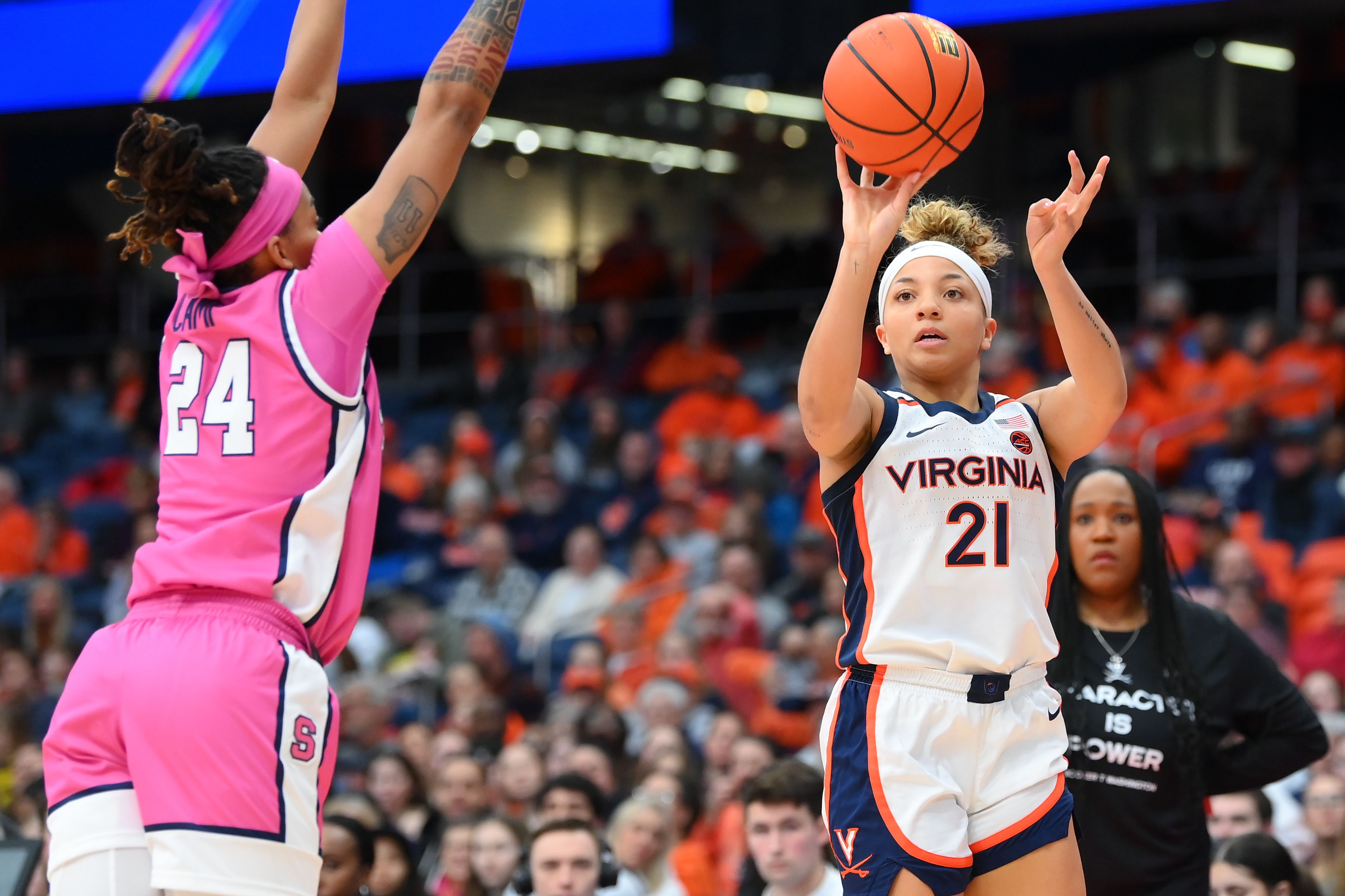 Virginia player in white jersey #21 shoots as a pink-clad defender #24 contests; crowd in orange fills the arena.