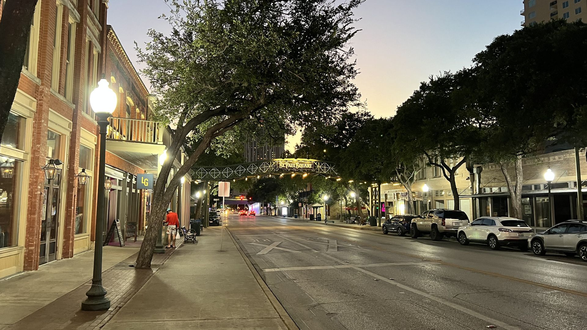 Urban street at dusk: red brick storefronts on the left, vintage lamps, a leaning tree center, an arch reading St. Paul Square, parked cars on the right, and a tall building against a blue-yellow sunset.