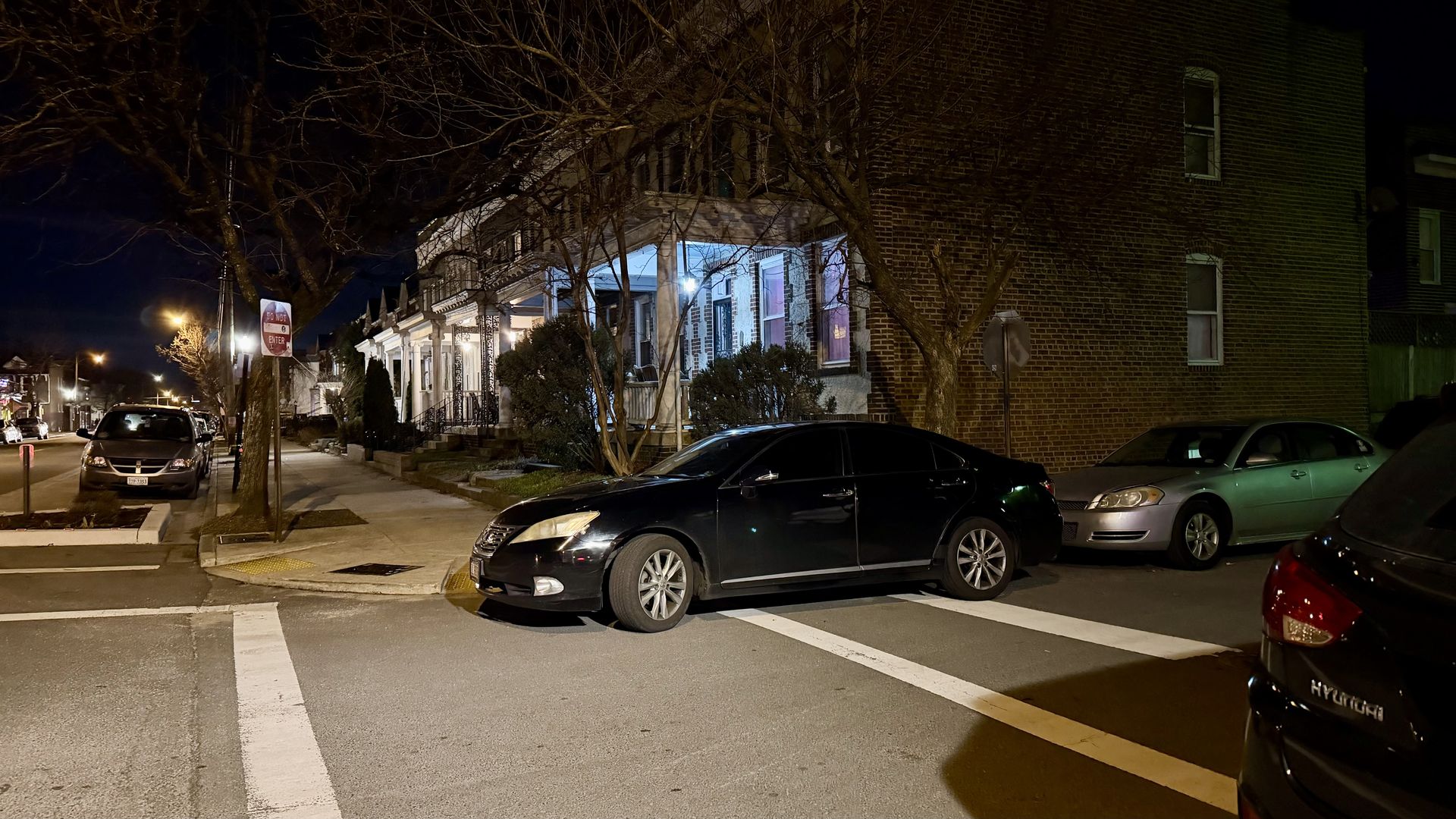Black sedan parked across a street intersection at night near a brick building and porch-lit houses with leafless trees lining the sidewalk under streetlights.