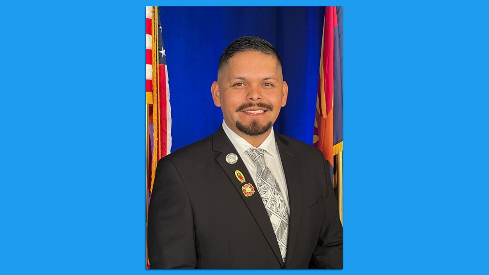 Smiling man in a black suit, white shirt, and patterned gray tie, wearing three lapel pins, posed in front of American and Arizona flags with a blue backdrop.