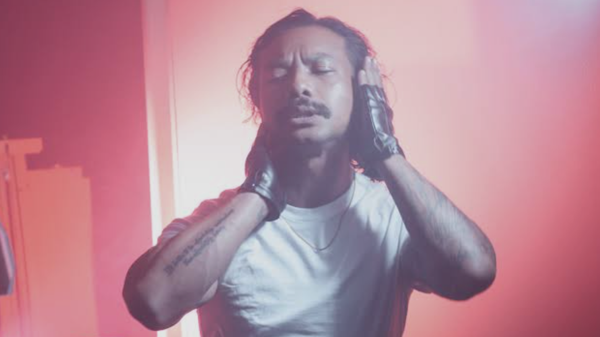 A man with dreadlocks in a white T-shirt and gloves stands on a pink-lit stage, eyes closed, hands pressed to the sides of his head, with a microphone stand visible in the foreground.