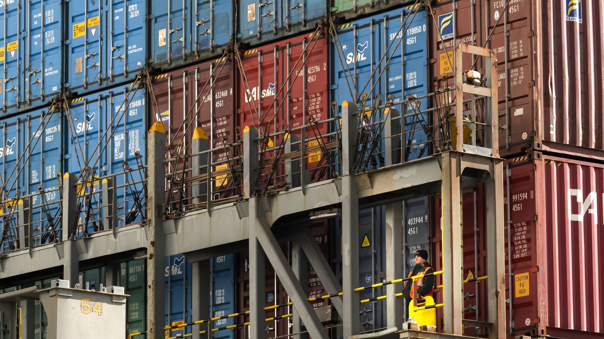 A worker in a safety vest and hard hat stands on a platform amid stacked shipping containers in red, blue, and green. Metal beams, railings, and cables secure the containers in an industrial port setting.