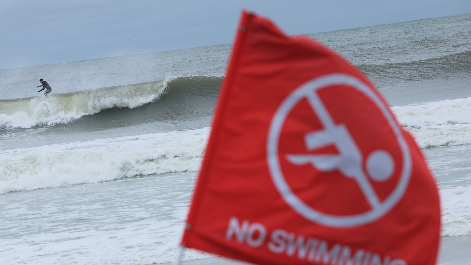 Red flag with a white no swimming symbol and text on a beach, waves crashing, and a surfer riding a wave in the background under a cloudy sky.