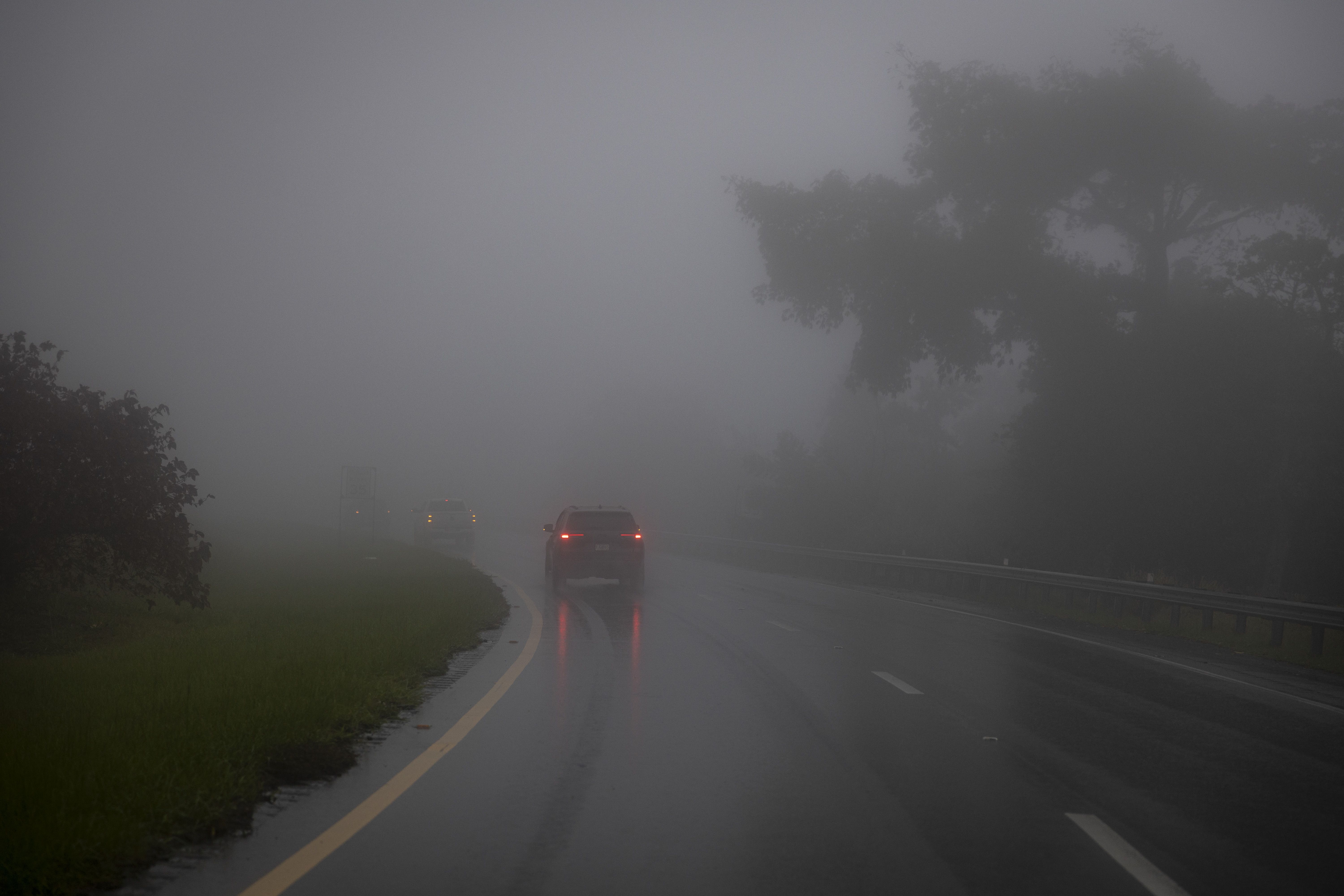Vehicles driving on a wet, foggy road with limited visibility, surrounded by faintly visible trees and greenery on a rainy day.