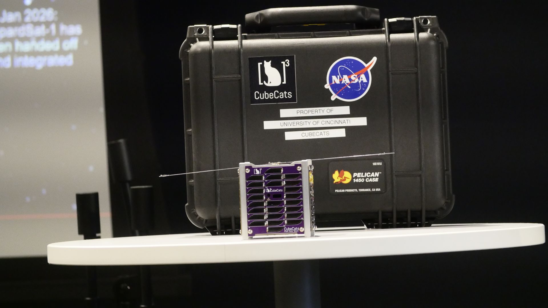 Black protective case on a white table, covered with stickers: CubeCats logo (white cat), NASA roundel, and a label reading PROPERTY OF UNIVERSITY OF CINCINNATI CUBECATS. The LEOPARDSat-1 sits in front.