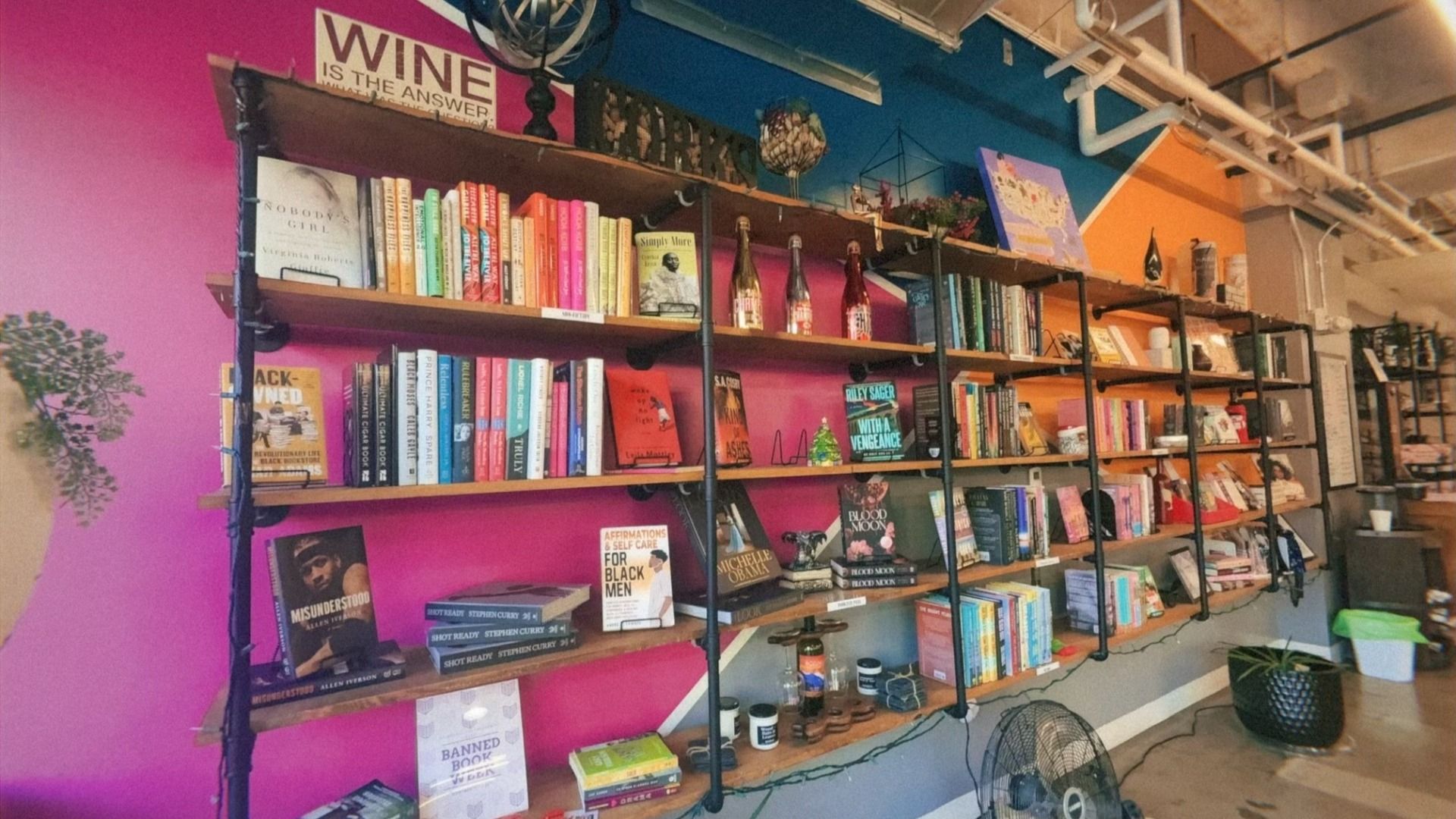 Bookshelves with diverse books, some bottles, decorative items, and a fan below. Walls painted pink, blue, and orange with a sign reading "WINE IS THE ANSWER."
