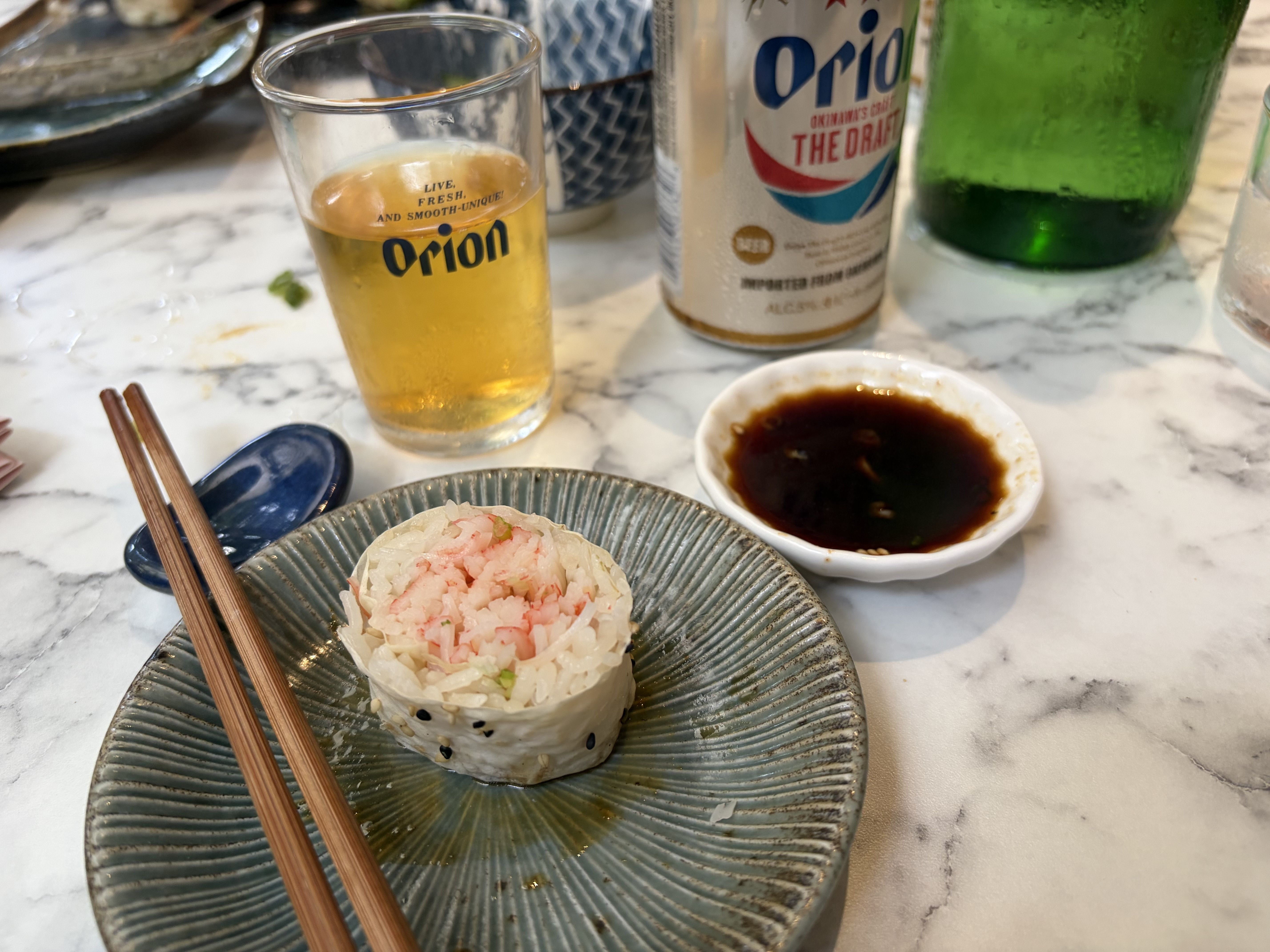 Close-up of a marble table: sushi rice cups on a blue plate with chopsticks, a soy sauce dish, and a glass and a can of Orion beer beside a green bottle in the background.